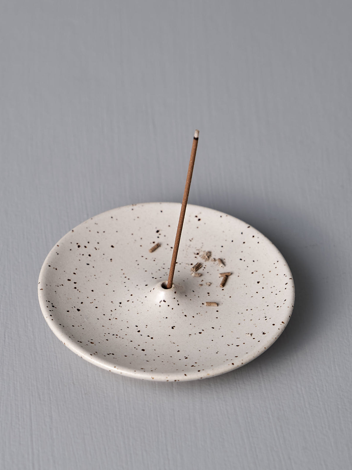 A single burning incense stick is placed in Nicola Shuttleworth&#39;s handmade Speckled White Incense Holder on a plain grey surface.