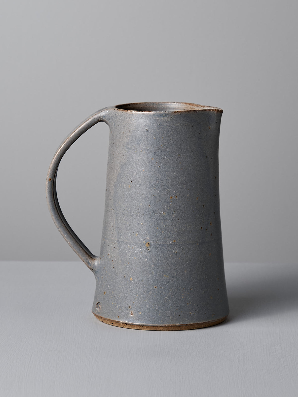 The &quot;Jug – Dusty Blue&quot; by Nicola Shuttleworth, a grey ceramic water jug with a handle and a stunning blue satin glaze, sits elegantly on a light grey surface against a plain grey background. This exquisite piece exemplifies the allure of handmade products, blending craftsmanship with functional art.