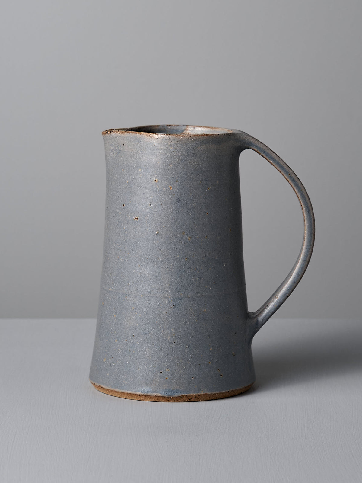 A Nicola Shuttleworth Jug – Dusty Blue, handmade with a handle and slight spout, sits on a gray surface against a matching gray background. This weighted water jug features a satin glaze interior, adding elegance to its functional design.