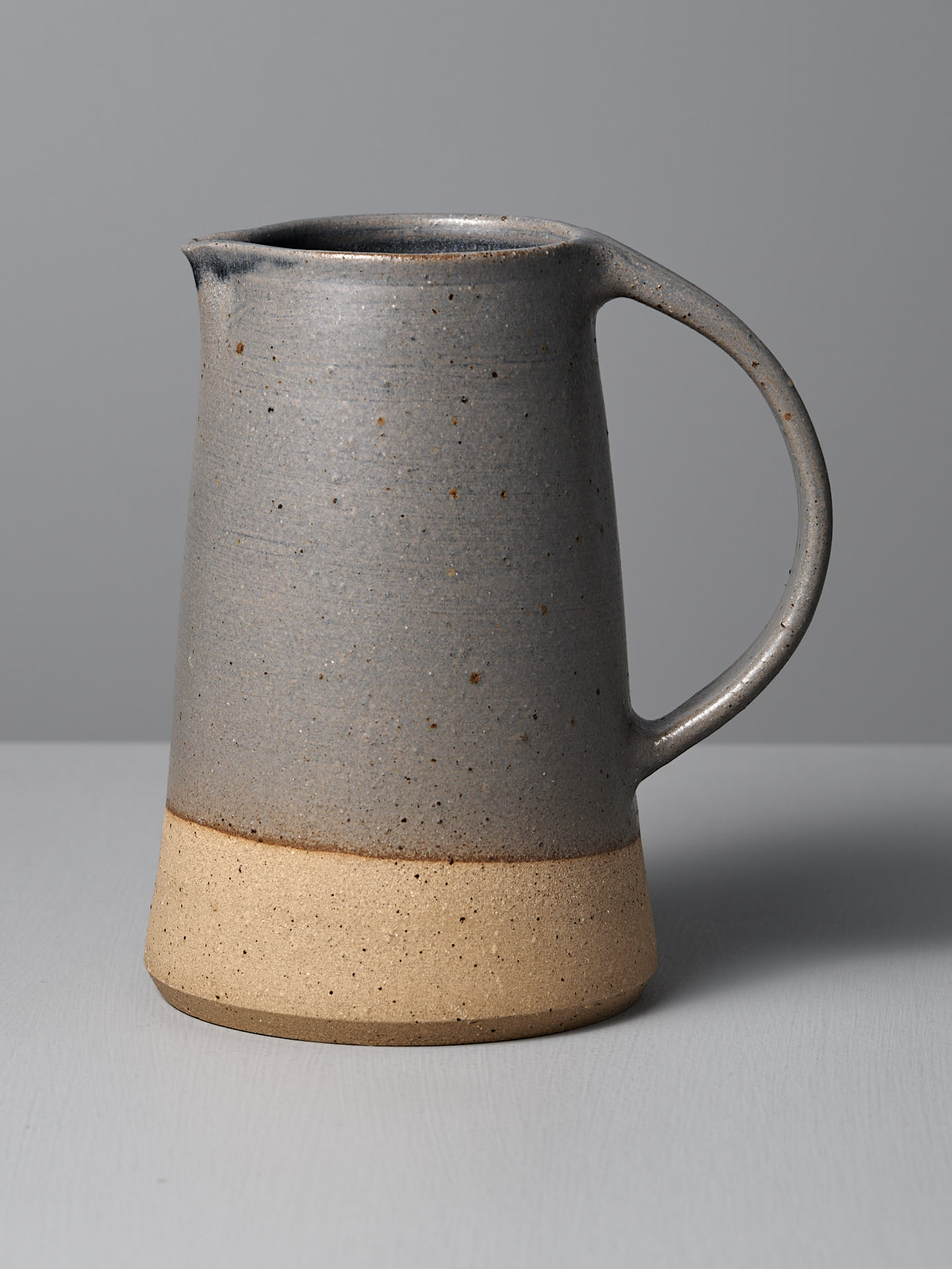 The Nicola Shuttleworth Jug – Dusty Blue, handmade with a semi-translucent blue glaze on the upper half and a textured raw base, rests on a light gray surface against a plain gray background.