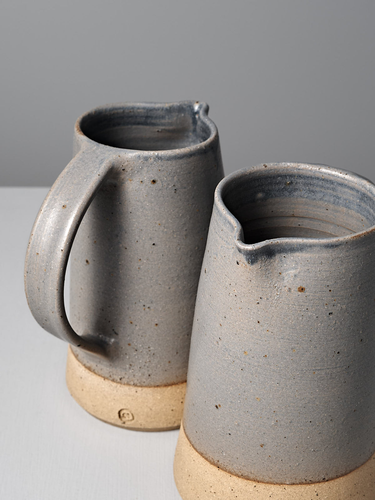 Two Nicola Shuttleworth Jug – Dusty Blue ceramic pitchers with a textured raw base and dusty blue speckled glaze are placed side by side on a light surface against a neutral background.
