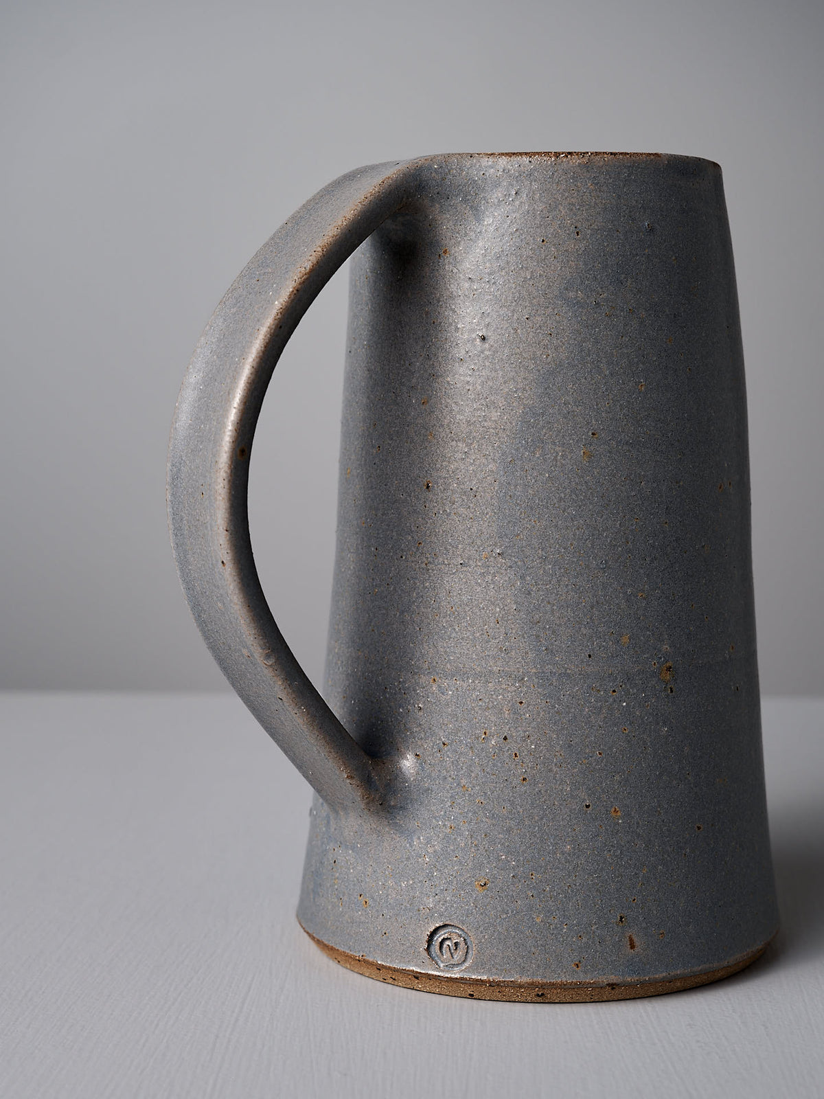 A Dusty Blue ceramic pitcher by Nicola Shuttleworth, with a textured finish and a single, large handle, featuring a satin glaze interior, placed on a light gray surface against a light gray background.