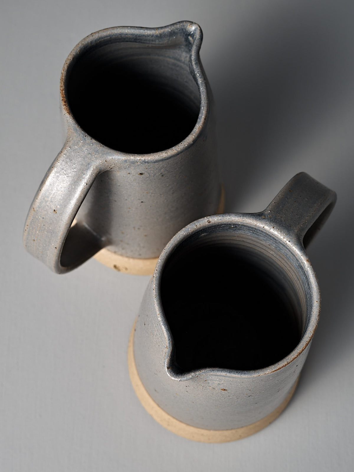 Two Nicola Shuttleworth Jug – Dusty Blue ceramic pitchers with textured raw bases and semi-translucent blue glaze are positioned side by side on a light surface, viewed from above.