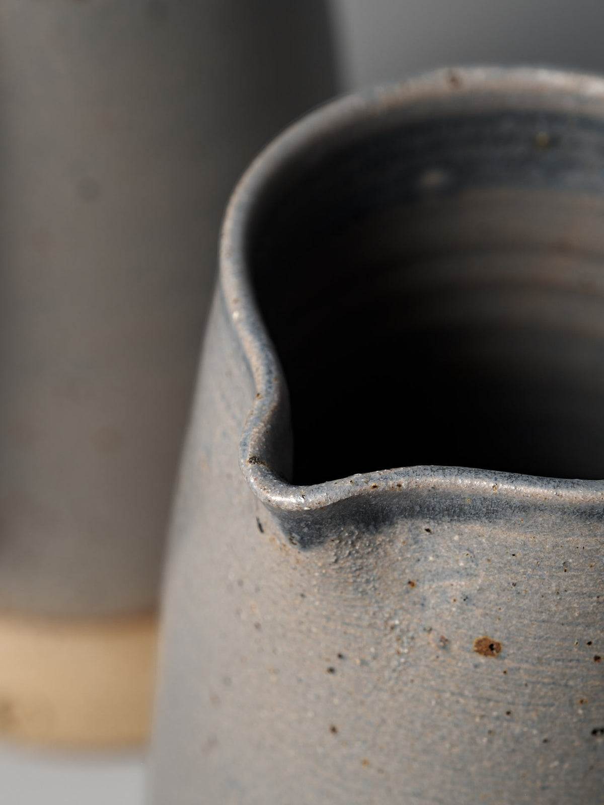 Close-up of Nicola Shuttleworth’s Jug – Dusty Blue, a handmade piece with a small spout, semi-translucent blue glaze, and textured raw base. A similar vessel appears softly blurred in the background.