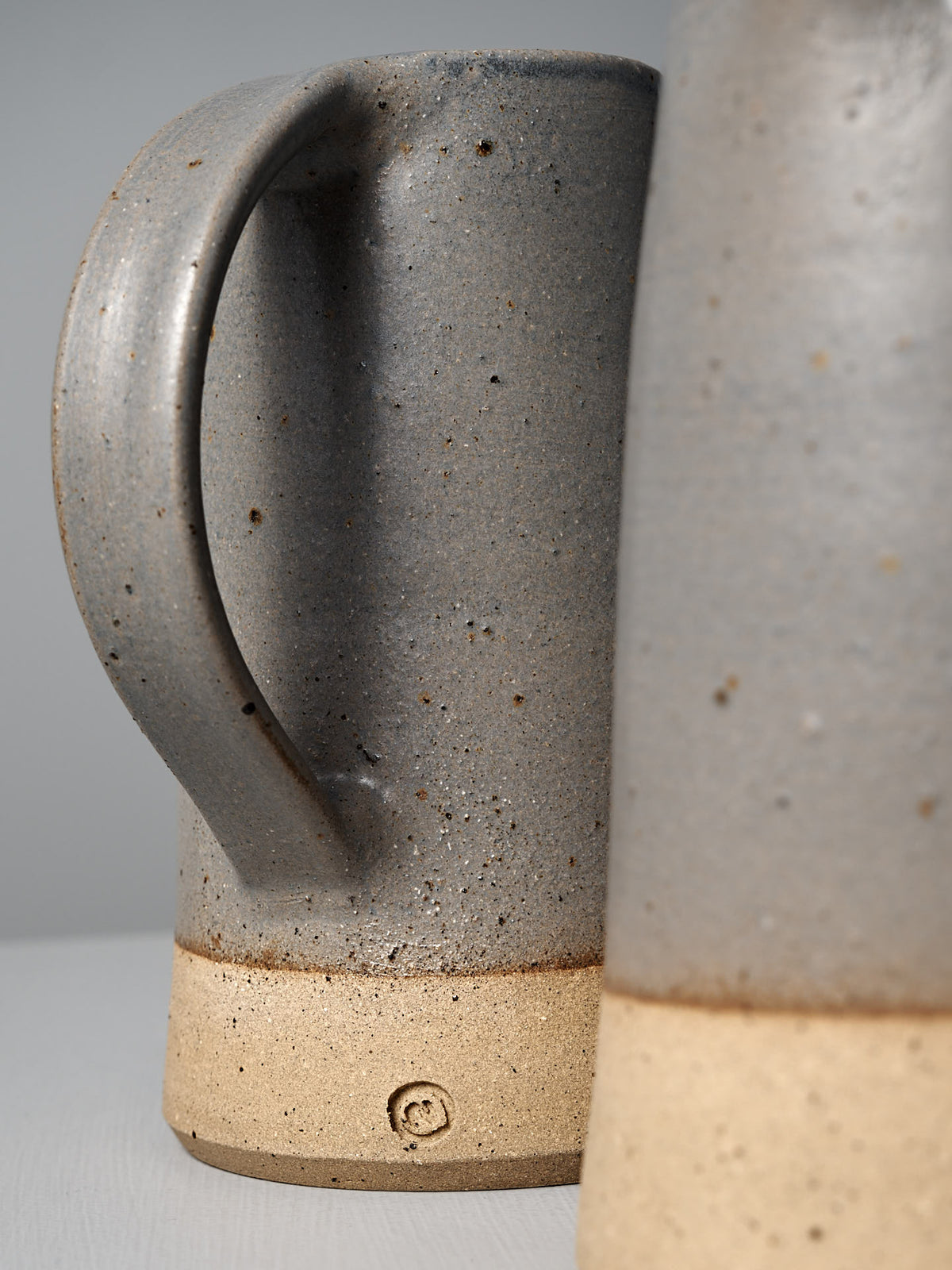 Close-up of the Nicola Shuttleworth Jug – Dusty Blue, featuring handmade ceramic construction with a speckled gray and semi-translucent blue glaze, unglazed textured raw base, visible handle, and the maker&#39;s mark.