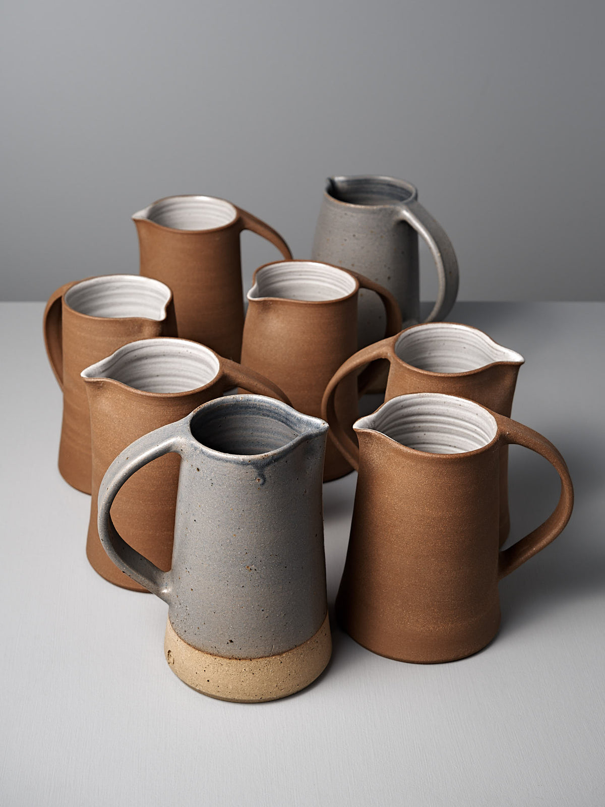 Seven ceramic pitchers, including Nicola Shuttleworth’s Jug – Dusty Blue with a handmade textured base, are arranged on a light surface against a plain gray background. Six are brown and one is blue.