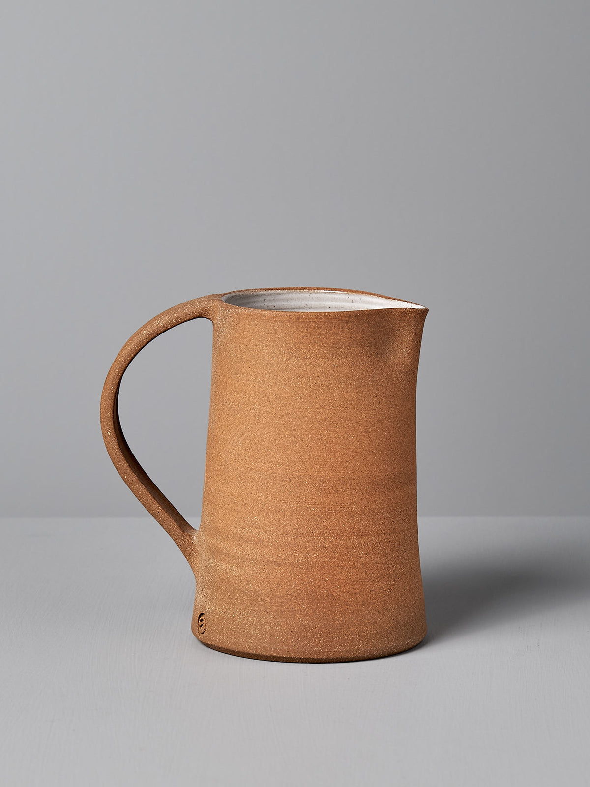 The Nicola Shuttleworth Jug – Textured, showcasing its intricate handmade design and practical handle, is set on a gray surface against a matching gray background.