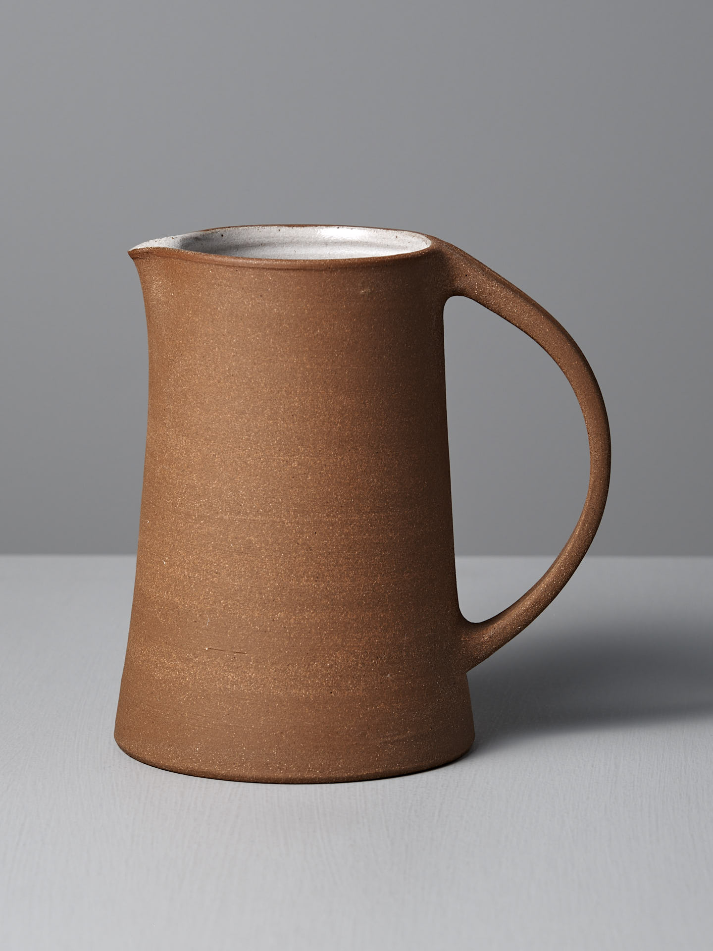 The Jug – Textured by Nicola Shuttleworth, handmade with a curved handle and raw unglazed brown exterior, rests on a light gray surface against a plain gray background.