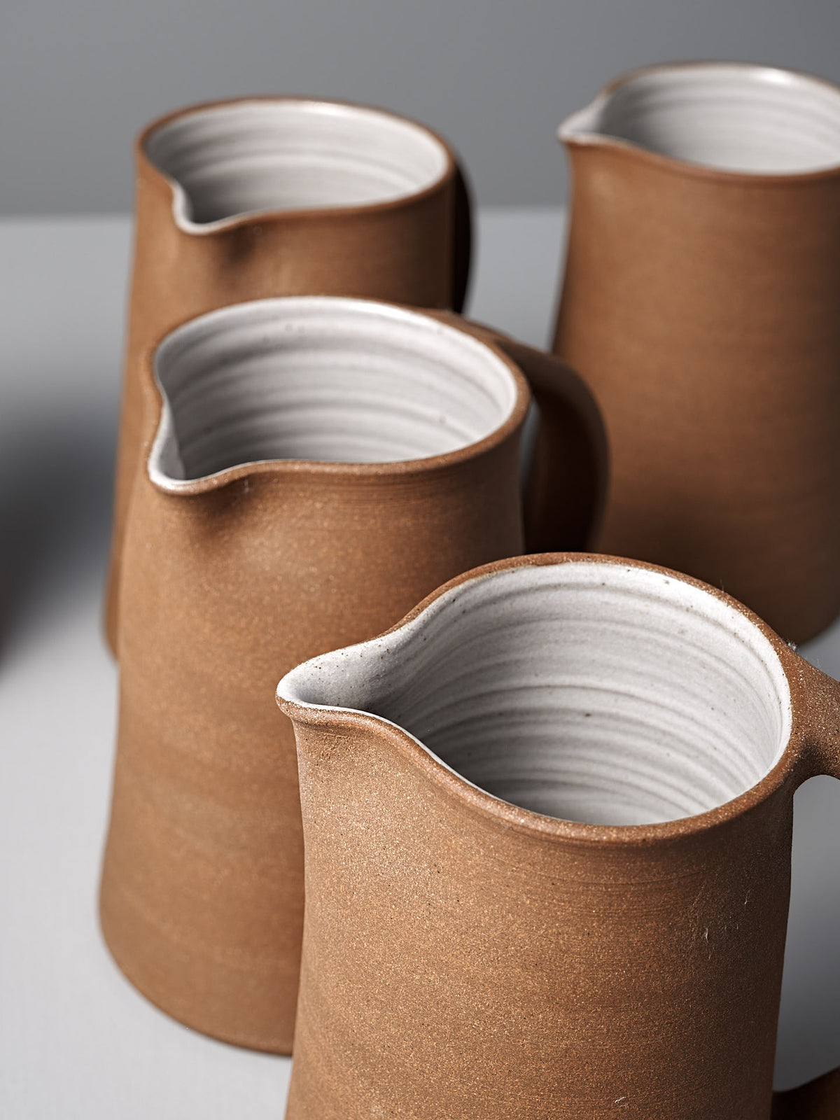 Four brown Nicola Shuttleworth &quot;Jug – Textured&quot; handmade jugs with white interiors sit on a light gray surface. Each features a simple matte finish, raw unglazed exterior, and wide spout.