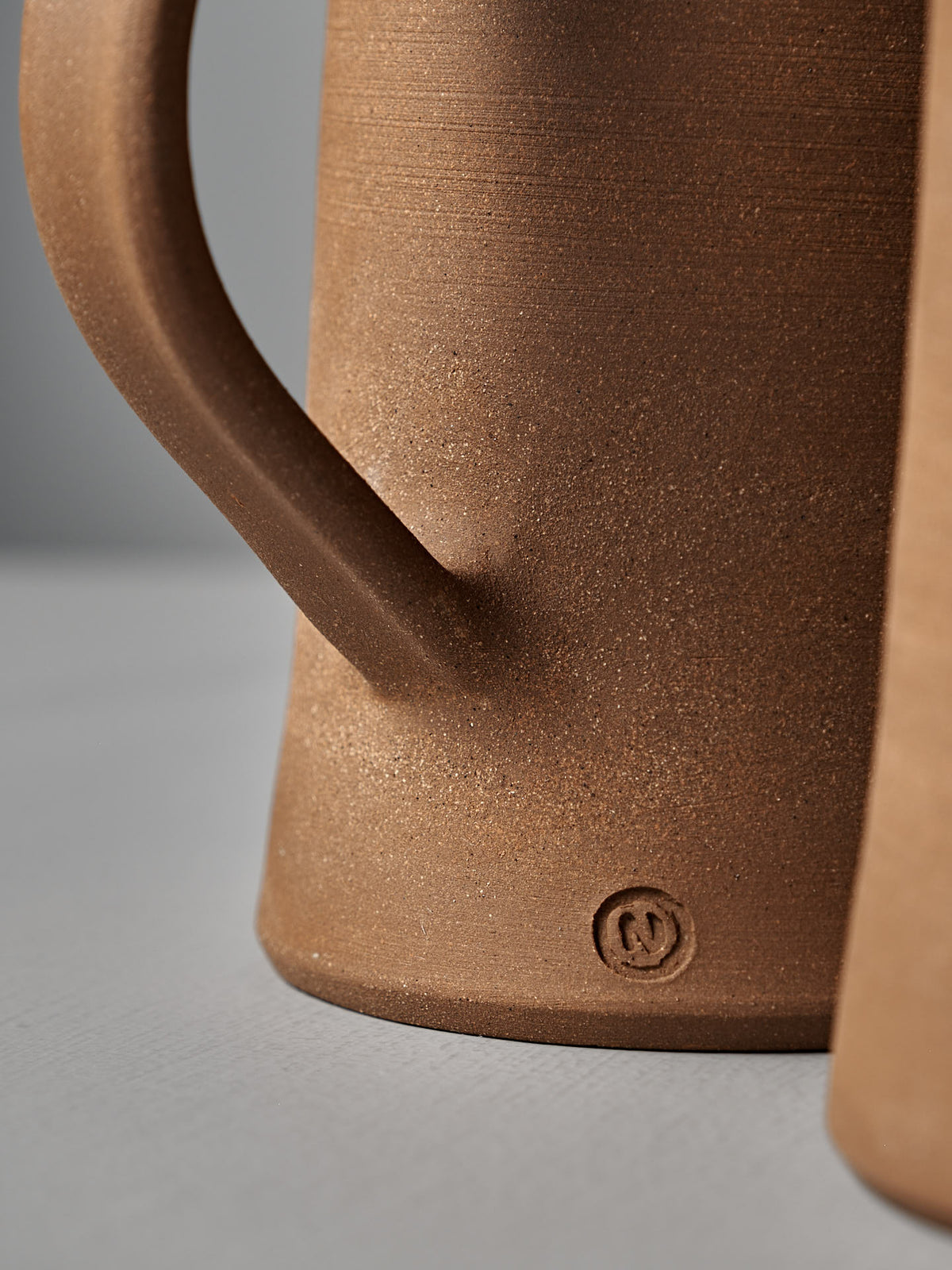 Close-up of the Jug – Textured by Nicola Shuttleworth, showcasing a brown satin glaze and a small maker’s mark near the base, set against a neutral background.