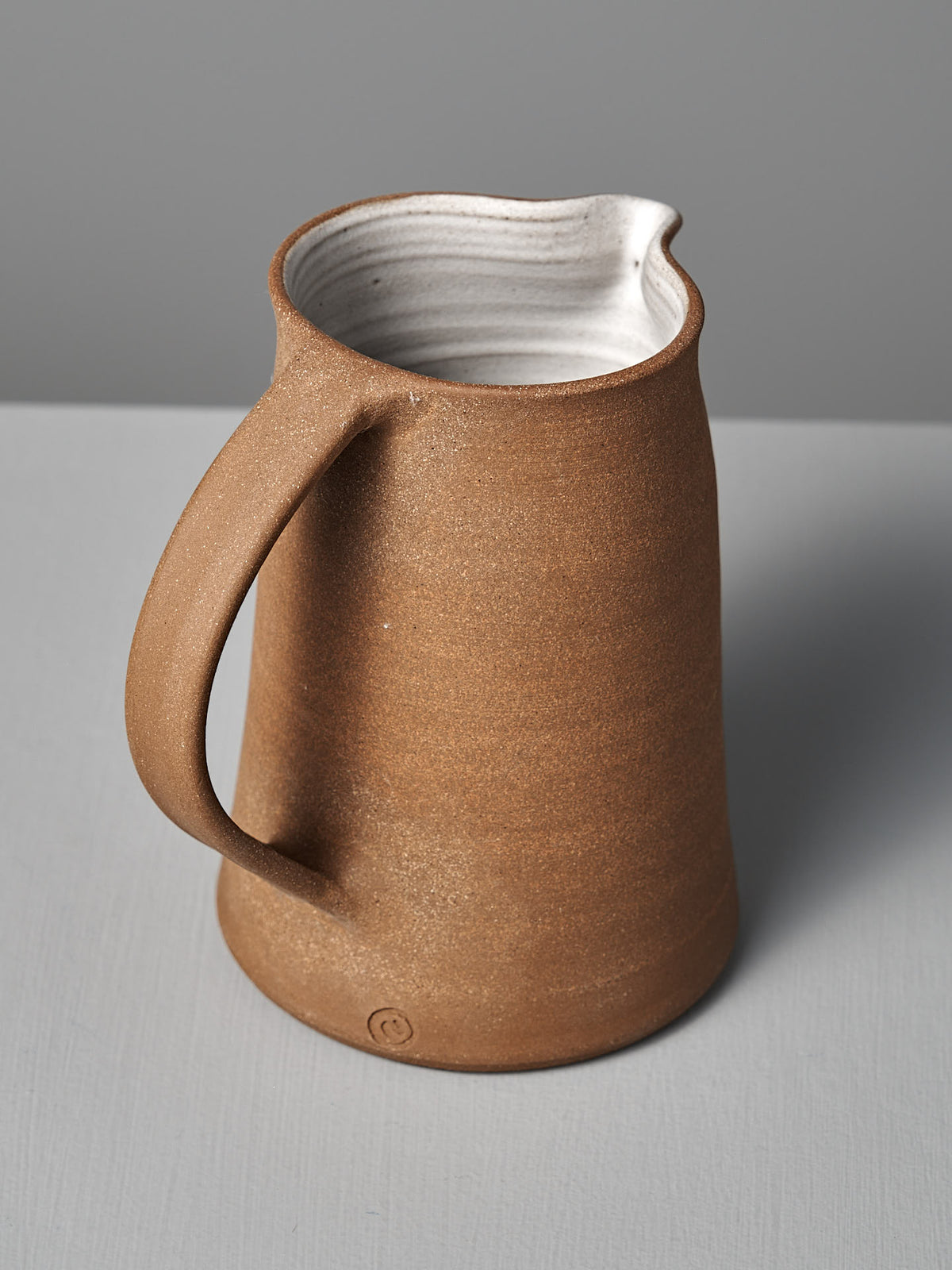 The Jug – Textured by Nicola Shuttleworth is a brown handmade piece with a curved handle and smooth white interior, featuring an unglazed exterior. It sits on a light gray surface with a gray background.