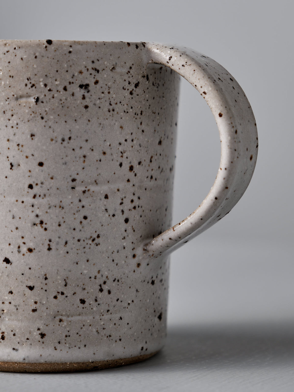 Close-up view of the Nicola Shuttleworth Mug – Speckled handle with a textured surface and grayish background, showcasing its speckled ceramic and warm white satin glaze.