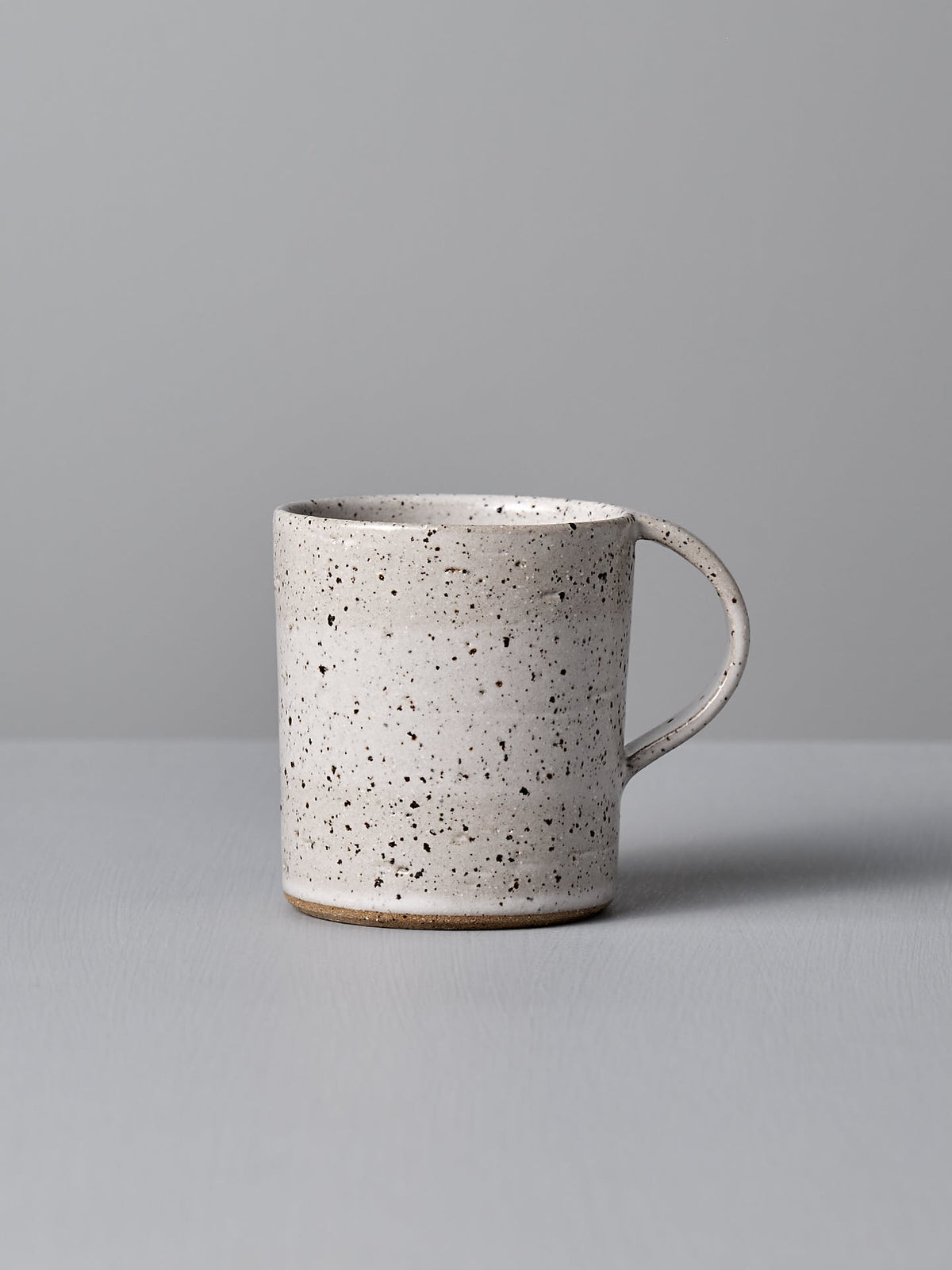 A Nicola Shuttleworth Mug – Speckled, featuring a handmade white ceramic design with black speckles, a handle, and a textured base, positioned against a light gray background.