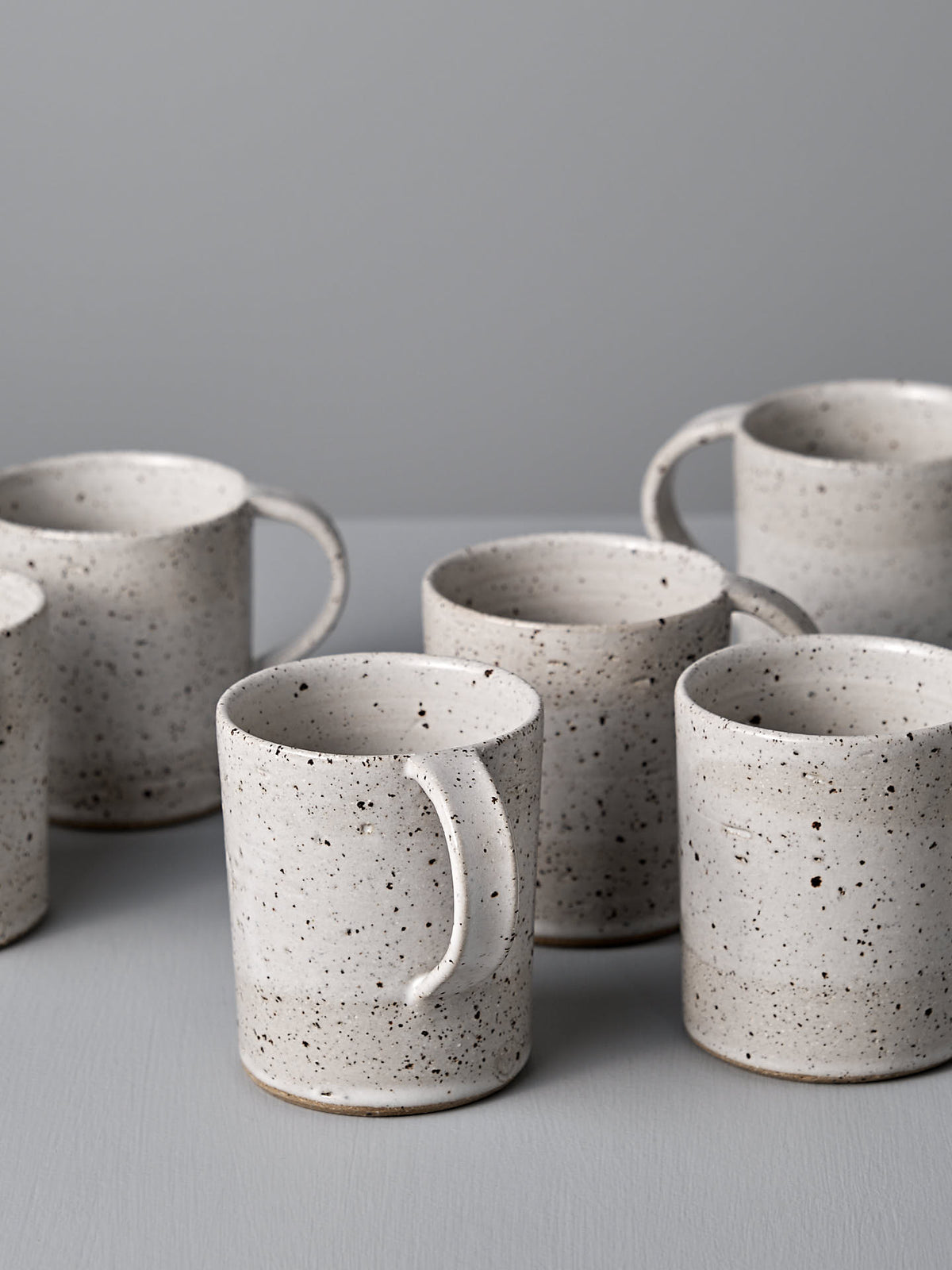 Six Nicola Shuttleworth &quot;Mug – Speckled&quot; handmade mugs with a satin glaze are arranged on a light grey surface against a light grey background.