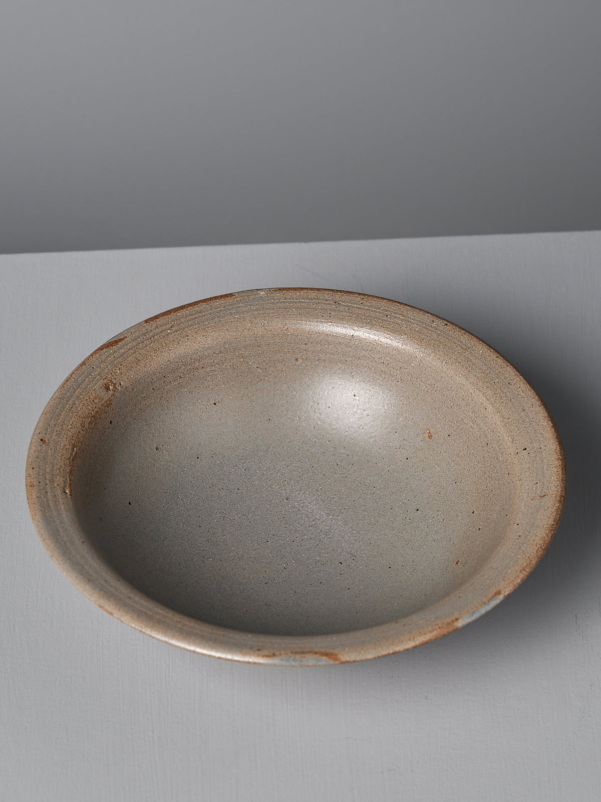 A serving bowl from Nicola Shuttleworth, named Blue Ash, sits on a light gray background. This beige ceramic bowl features a slightly textured surface and a blue ash satin glaze. The handmade stoneware item, crafted in Pōneke Wellington, boasts minor imperfections and subtle color variations around the edges.