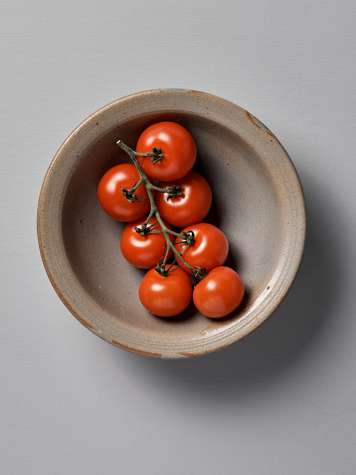 A cluster of red tomatoes on the vine sits in a round, beige ceramic Serving Bowl – Blue Ash by Nicola Shuttleworth, against a light gray background.