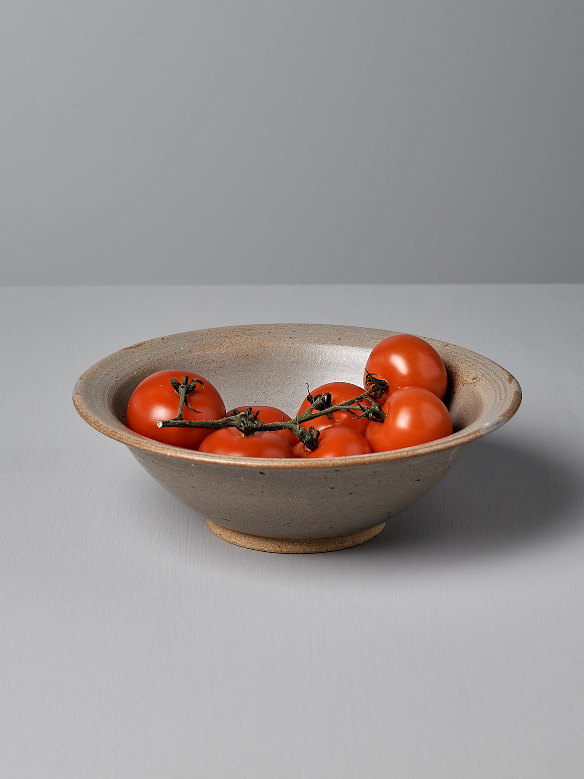 A Serving Bowl – Blue Ash by Nicola Shuttleworth from Pōneke Wellington, adorned with a blue ash satin glaze, cradles a bunch of ripe red tomatoes on the vine, all set against a light gray surface.