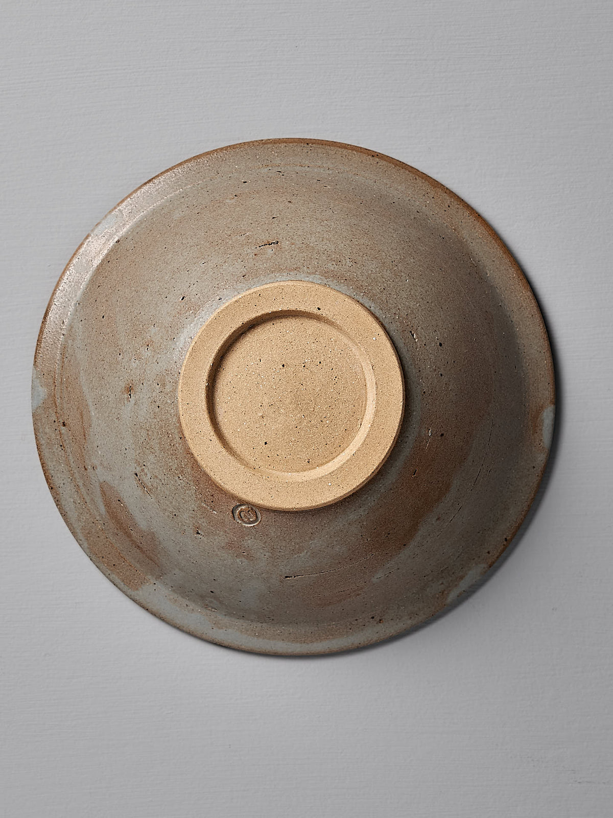 Top view of an upside-down handmade stoneware Serving Bowl – Blue Ash by Nicola Shuttleworth, featuring a beige and brown speckled glaze, resting on a neutral gray surface. Crafted in Pōneke Wellington, its rustic charm is reminiscent of the blue ash satin glaze.