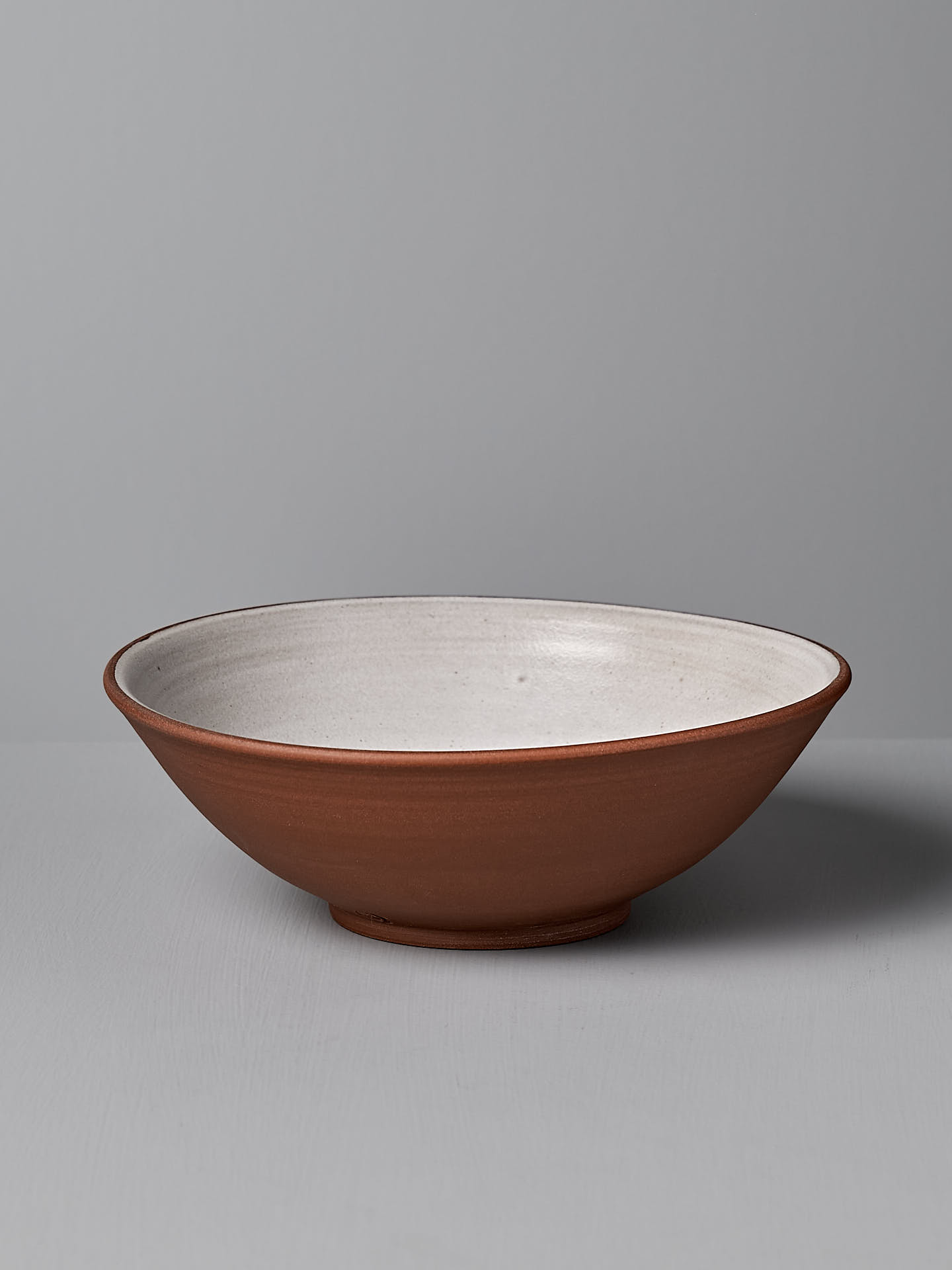 The Serving Bowl – Red Clay by Nicola Shuttleworth, a shallow ceramic pasta bowl with a white interior and brown exterior, adorned with a subtle blue ash satin glaze, sits on a flat surface against a gray background.