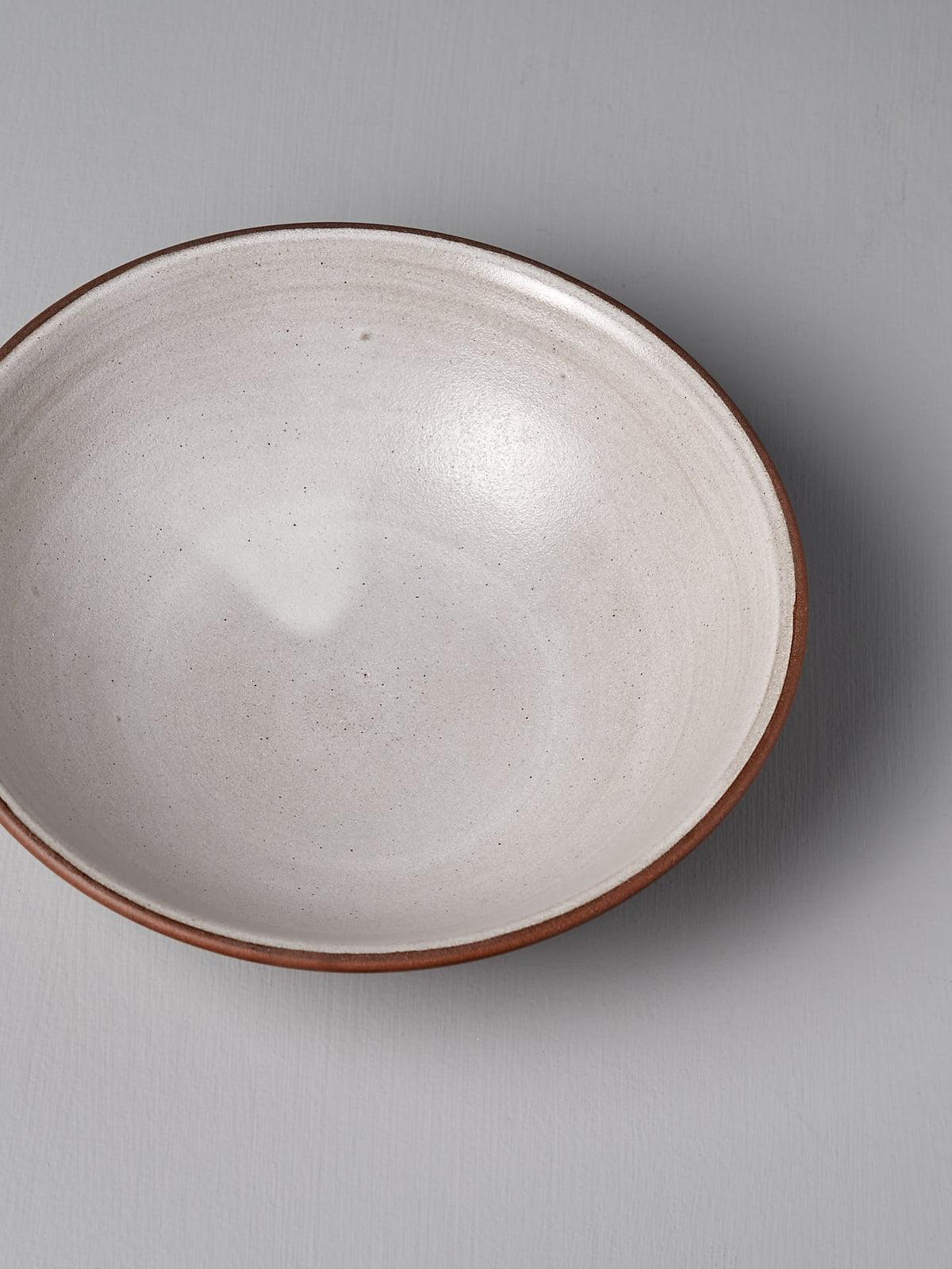 A handmade red clay serving bowl by Nicola Shuttleworth, featuring a smooth, light interior and a darker blue ash satin glaze exterior, rests on a flat surface.