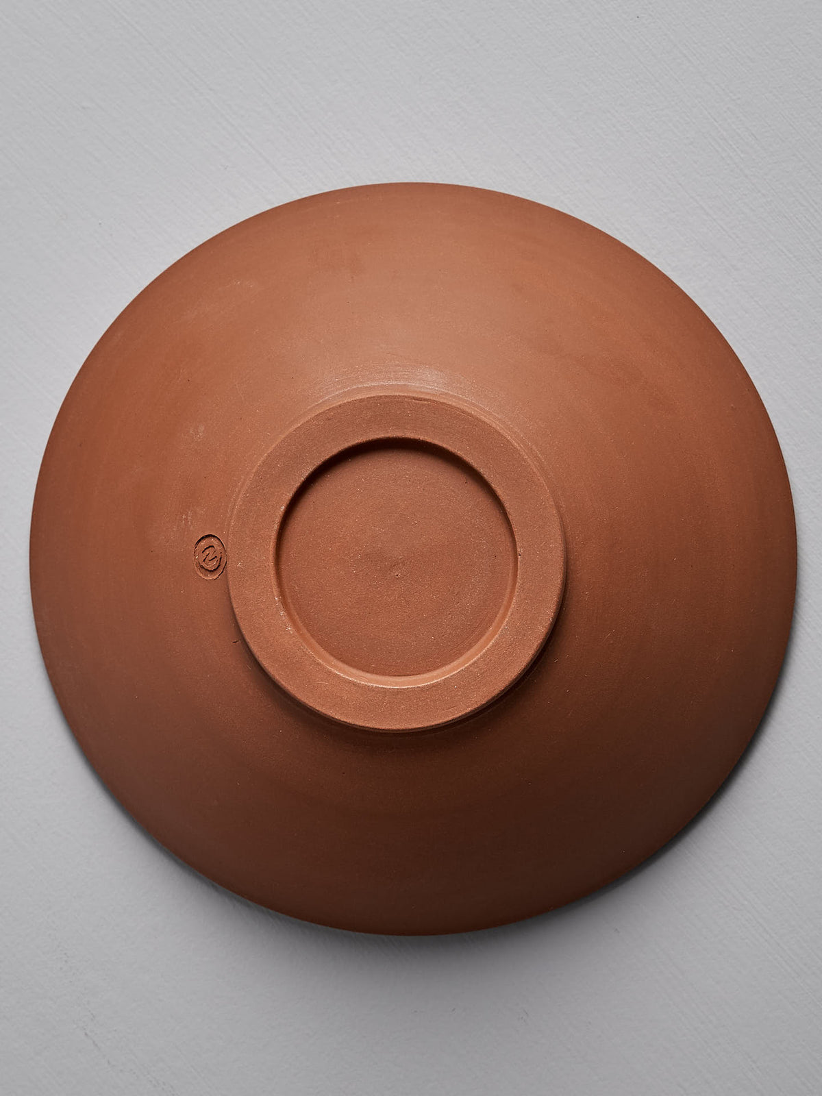 A Nicola Shuttleworth Serving Bowl – Red Clay is shown upside down on a plain, light gray surface, showcasing its handmade stoneware craftsmanship.