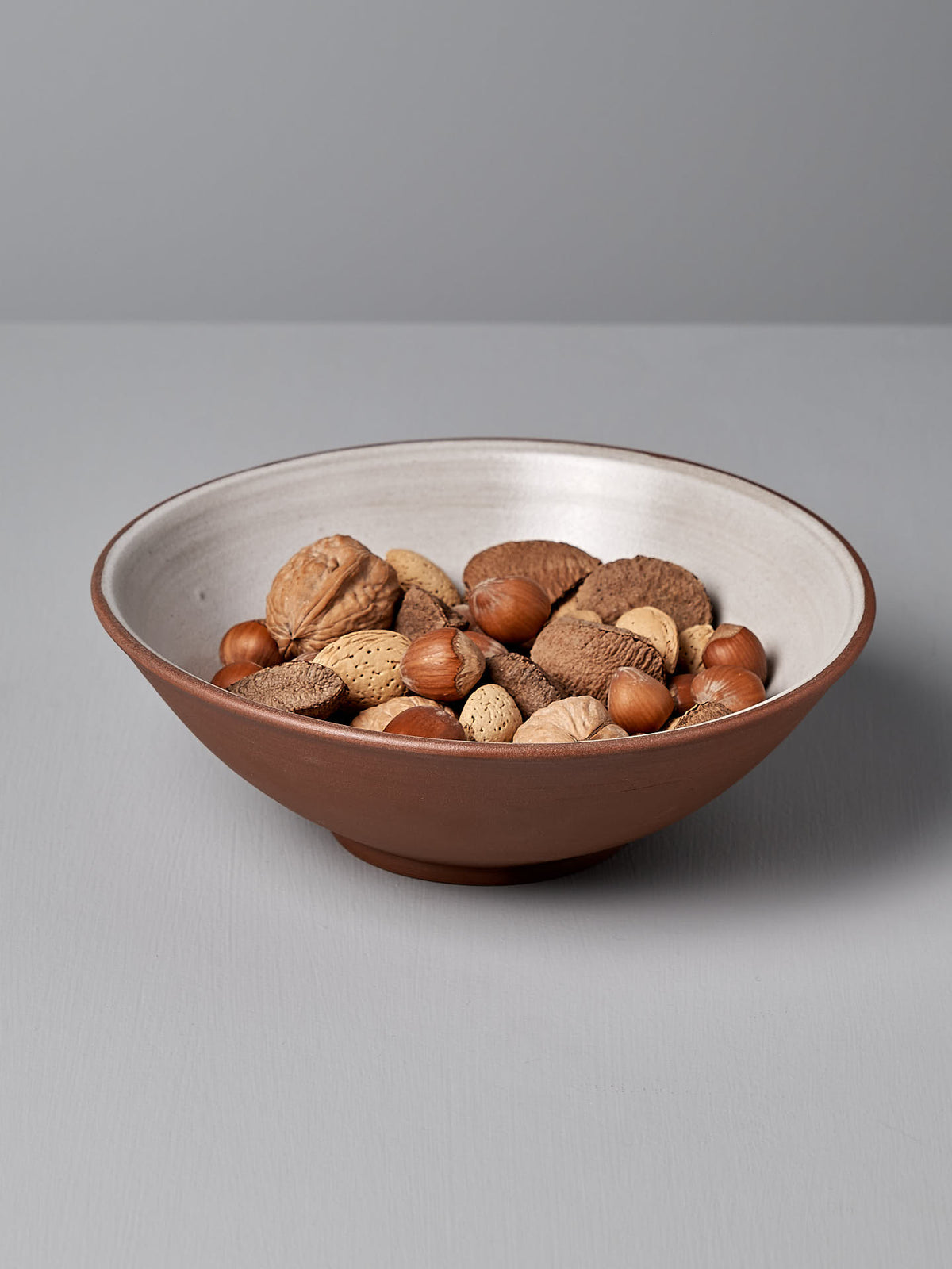 A Nicola Shuttleworth Serving Bowl – Red Clay, handmade from stoneware, filled with a variety of unshelled walnuts, hazelnuts, almonds, and pecans, placed on a gray surface.