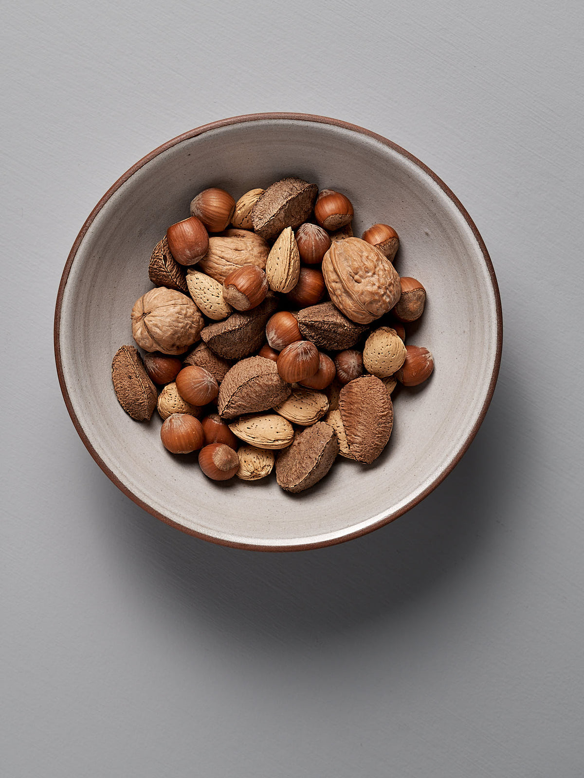 A white ceramic bowl, reminiscent of a pasta serving bowl, holds an assortment of unshelled nuts—including almonds, hazelnuts, and walnuts—placed on a light gray surface. The blue ash satin glaze adds a touch of elegance to this handmade stoneware piece by Nicola Shuttleworth.