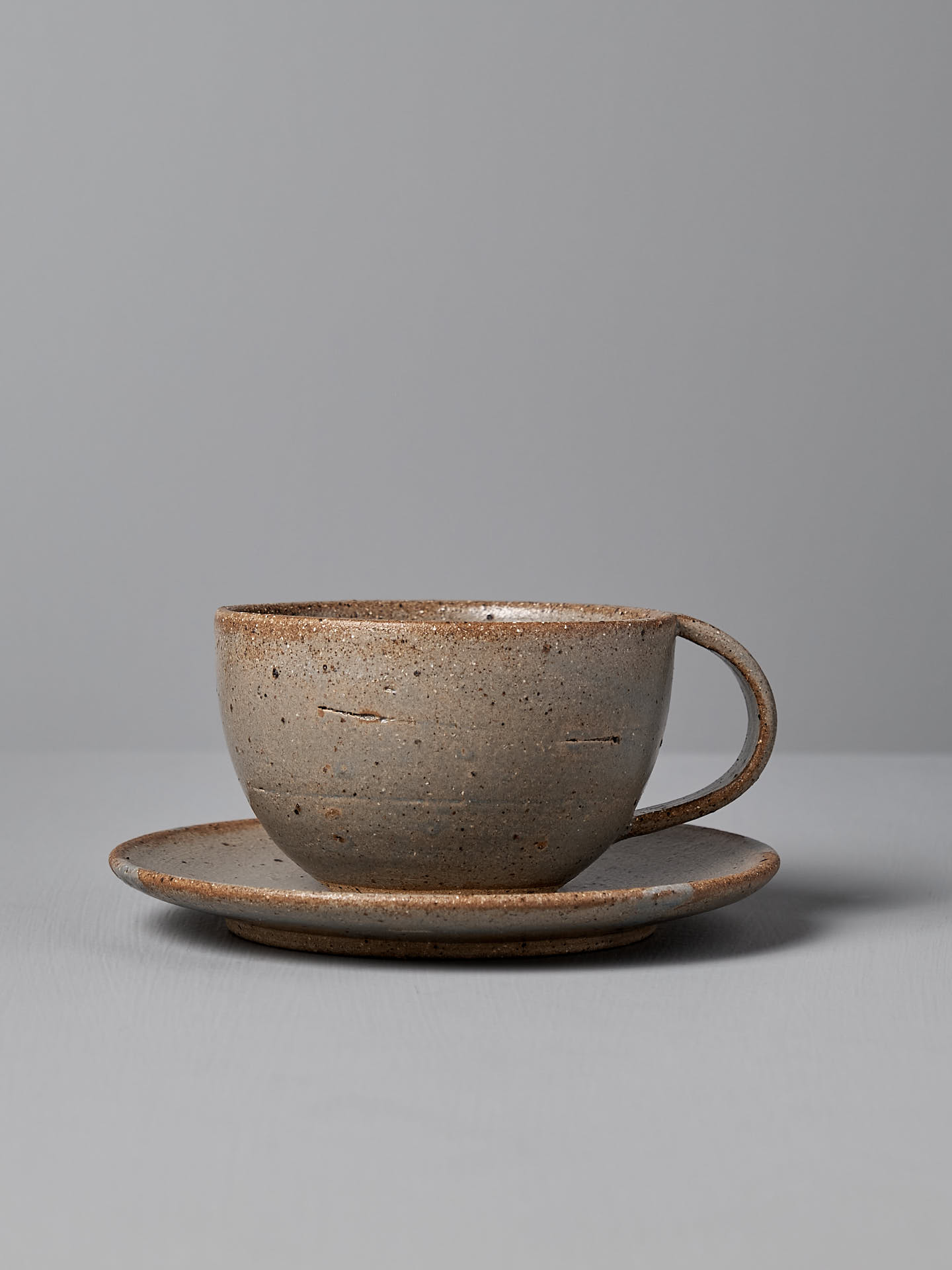 The Nicola Shuttleworth Cup & Saucer – Blue Ash, featuring a textured, handmade ceramic cup with a wide rim and handle on a matching saucer, adorned in a blue ash satin glaze, is set against a plain gray background.