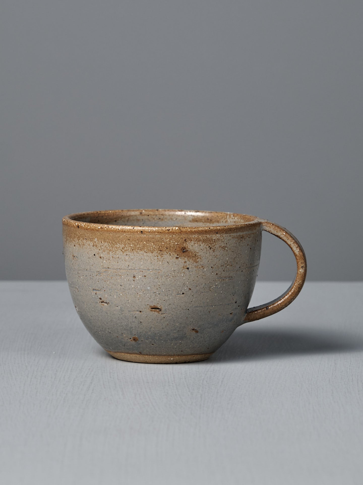 The Cup – Blue Ash features a handmade blue ash glaze with speckled gray and brown tones, displayed on a light gray surface against a plain gray background, showcasing the artisanal spirit of Wellington.