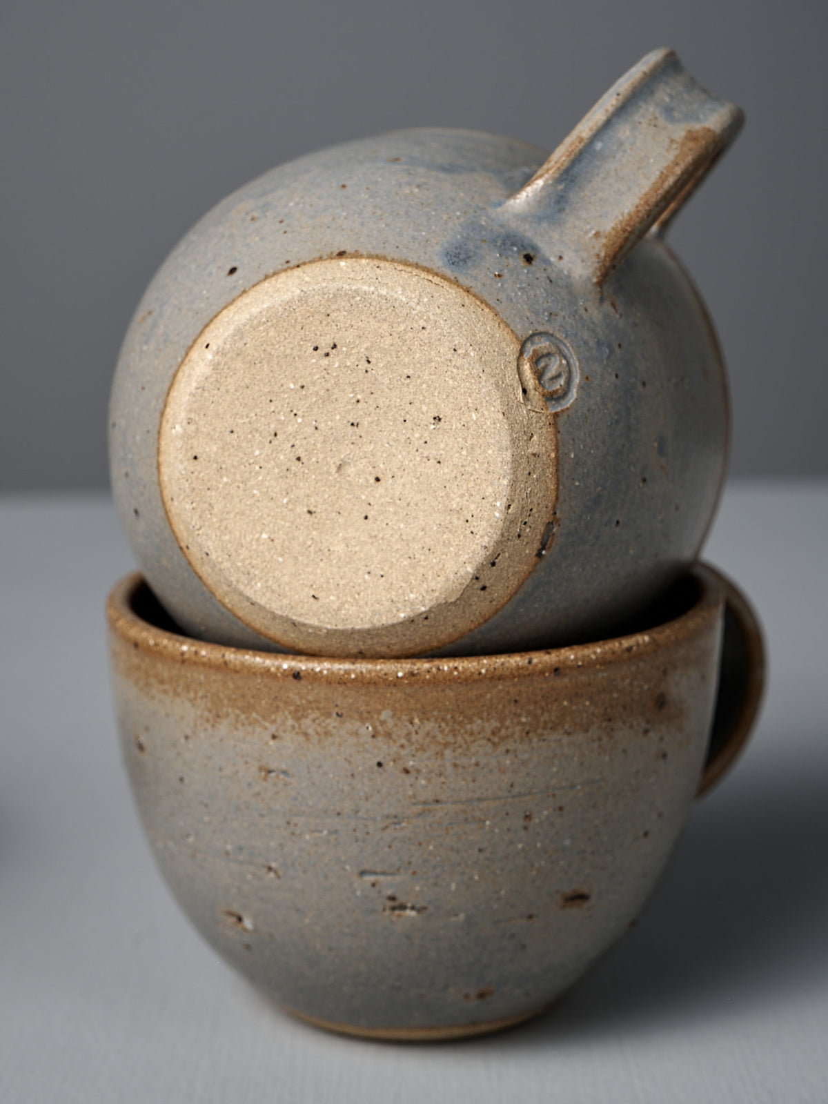 The Cup – Blue Ash features two handmade cups with speckled beige and gray glaze, blue ash accents, and visible handles, stacked together against a plain background. Inspired by Wellington craftsmanship.