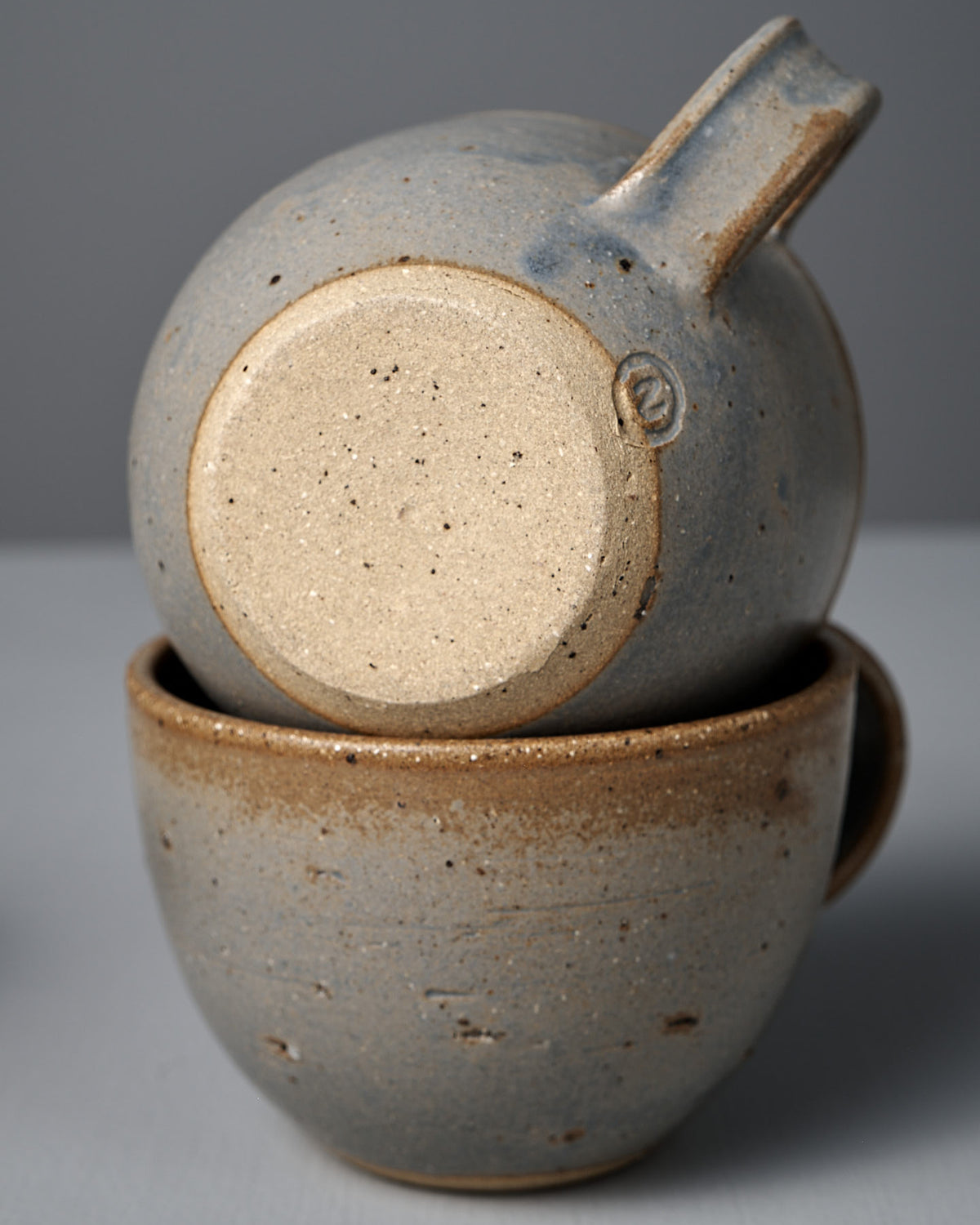 The Cup – Blue Ash features two handmade cups with speckled beige and gray glaze, blue ash accents, and visible handles, stacked together against a plain background. Inspired by Wellington craftsmanship.