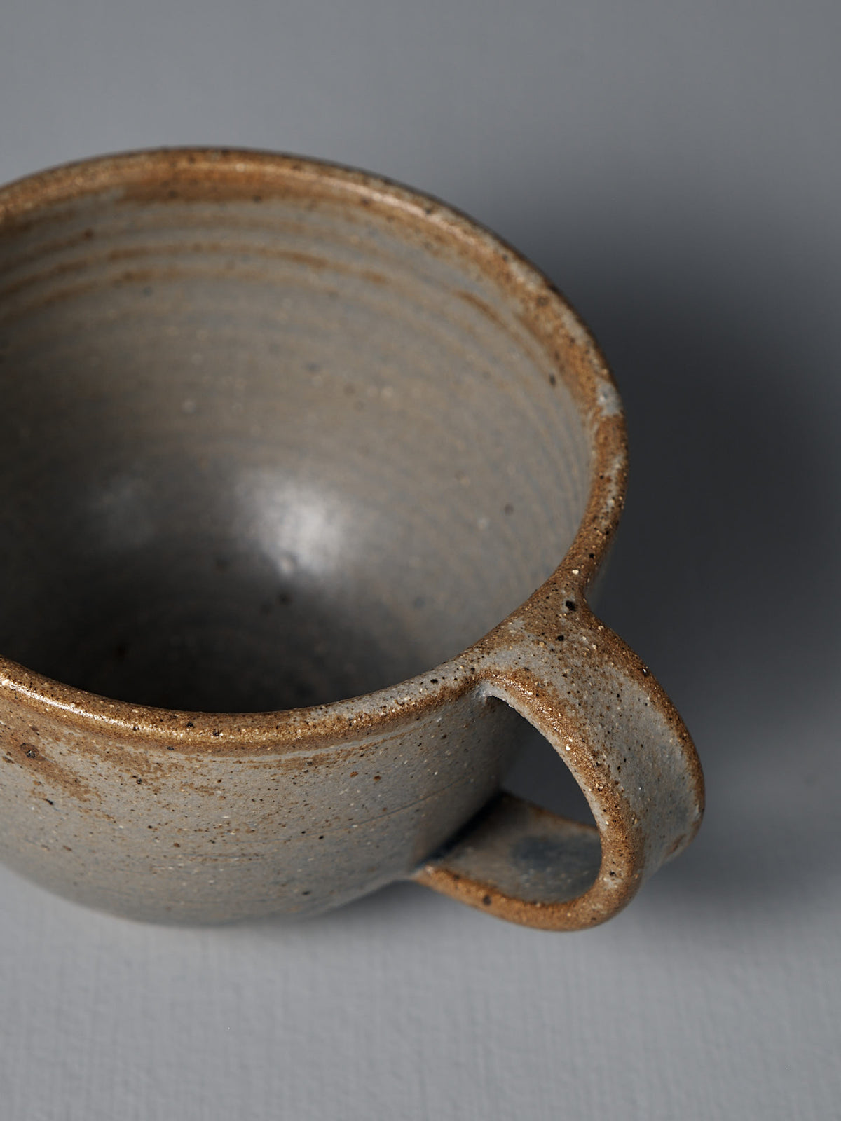 A close-up of the Cup – Blue Ash, a handmade ceramic with speckled brown and gray tones, shown on a plain light gray surface in Wellington.