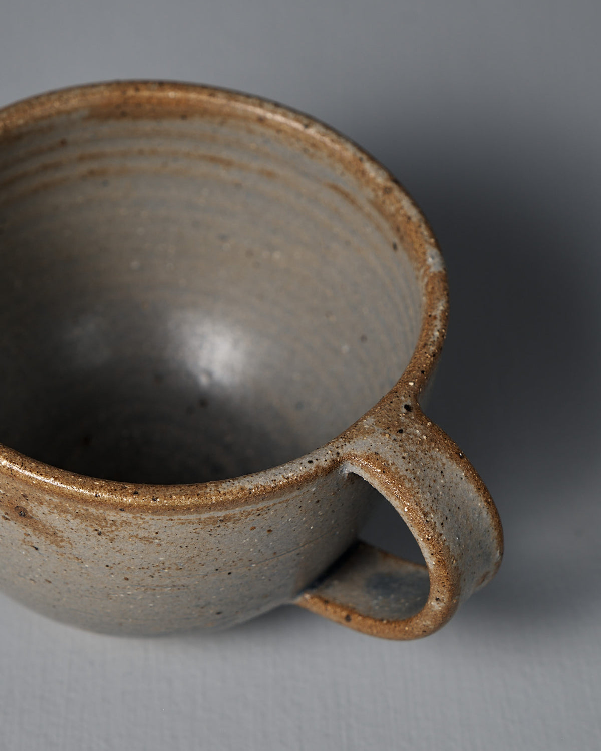 A close-up of the Cup – Blue Ash, a handmade ceramic with speckled brown and gray tones, shown on a plain light gray surface in Wellington.