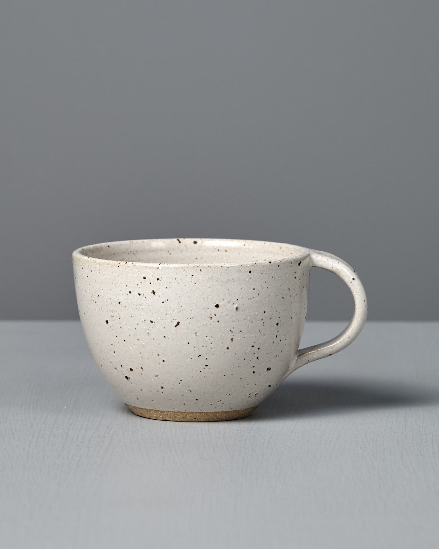 The Cup – Speckled White is a handmade, off-white cup with a handle and satin glaze, displayed on a light gray surface against a plain gray background.