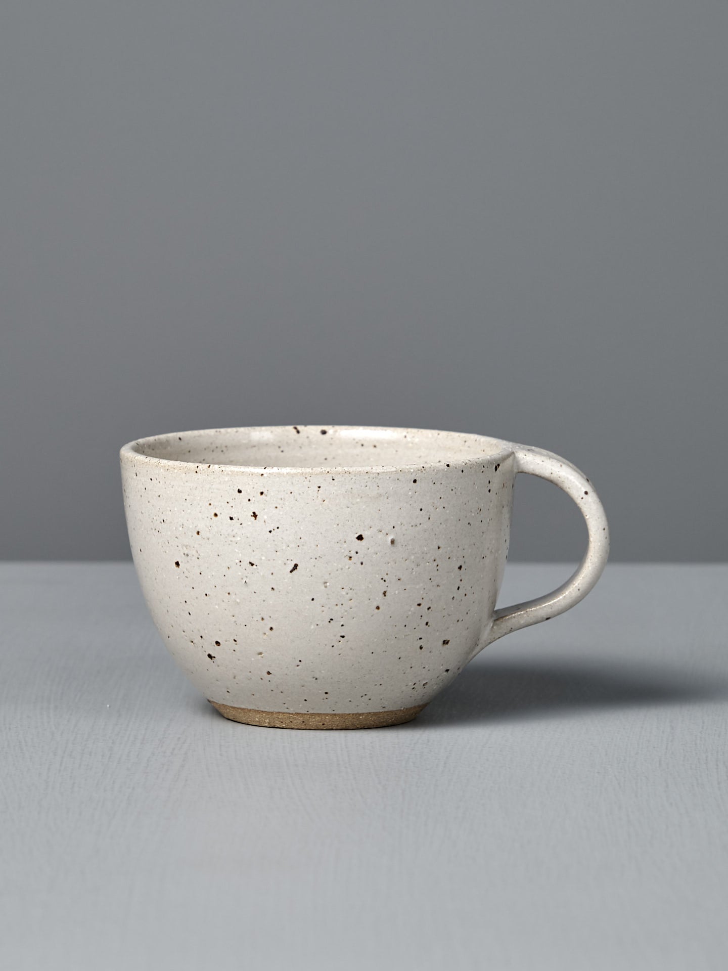 The Cup – Speckled White is a handmade, off-white cup with a handle and satin glaze, displayed on a light gray surface against a plain gray background.