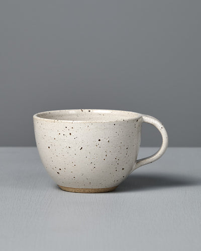 The Cup – Speckled White is a handmade, off-white cup with a handle and satin glaze, displayed on a light gray surface against a plain gray background.