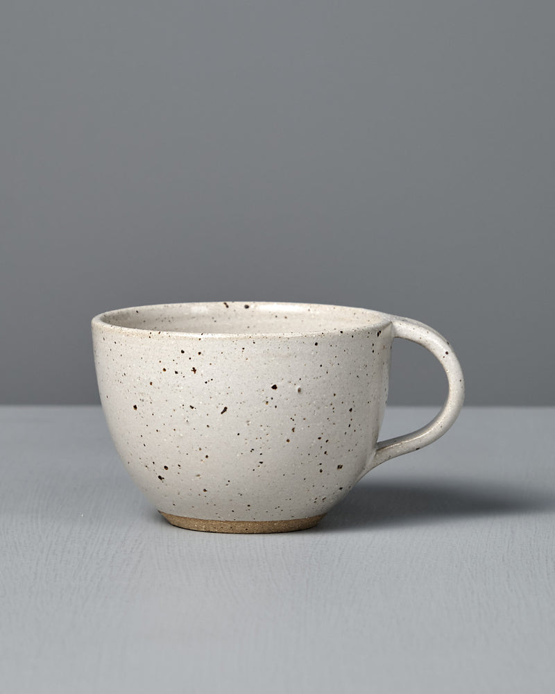 The Cup – Speckled White is a handmade, off-white cup with a handle and satin glaze, displayed on a light gray surface against a plain gray background.