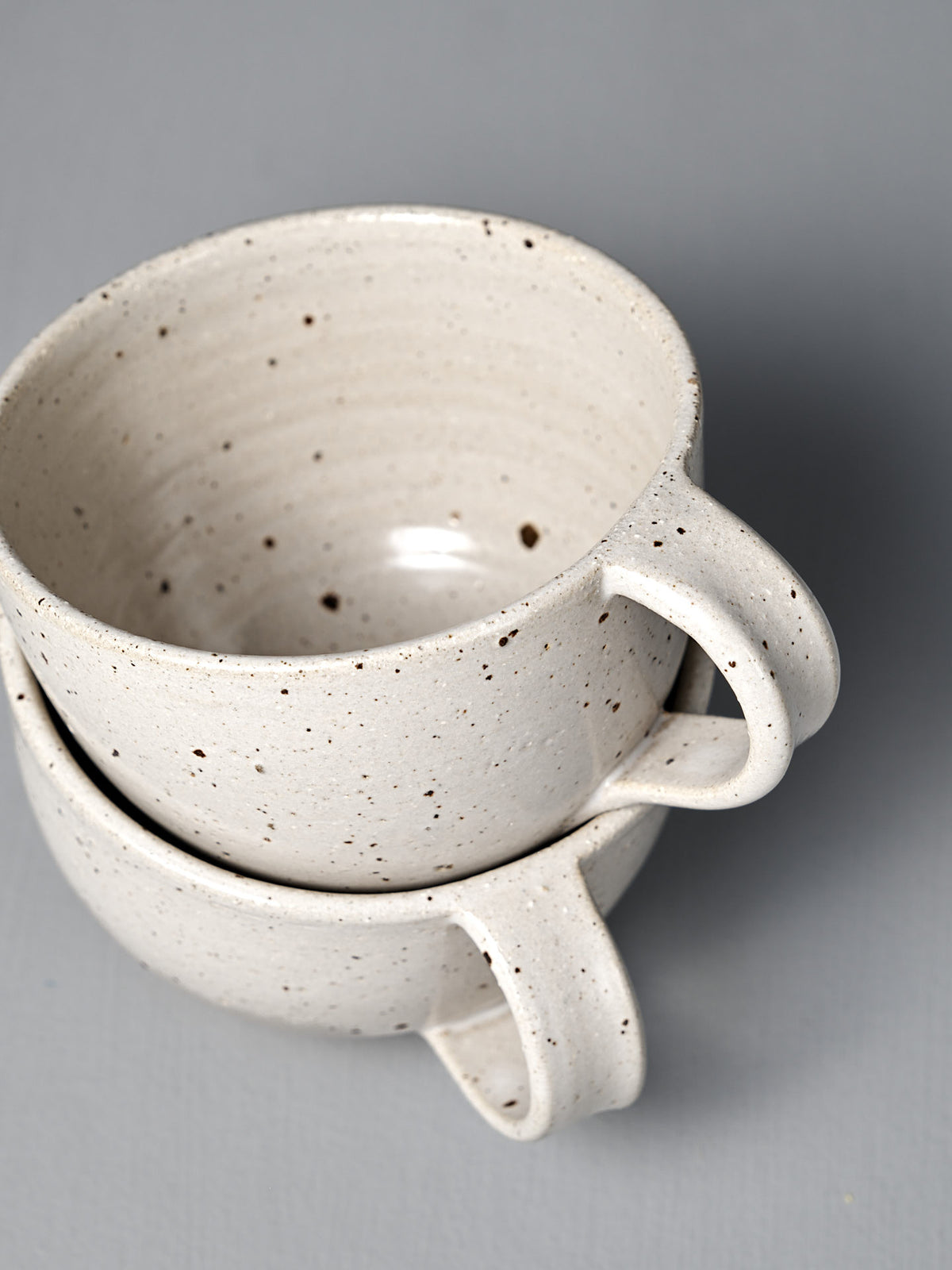 Two Speckled White handmade cups with a satin glaze are stacked together against a plain grey background, highlighting their artisanal craftsmanship.