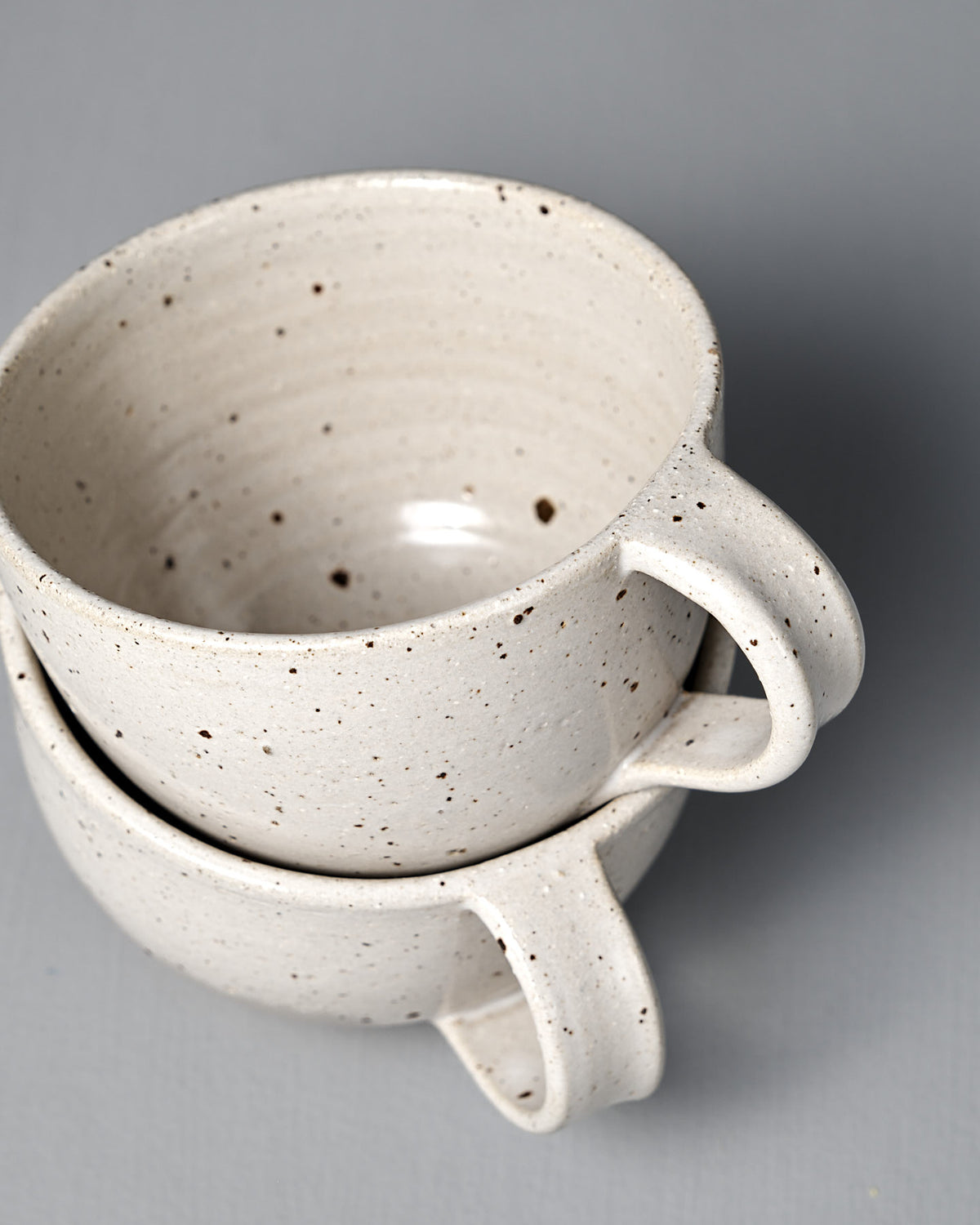 Two Speckled White handmade cups with a satin glaze are stacked together against a plain grey background, highlighting their artisanal craftsmanship.
