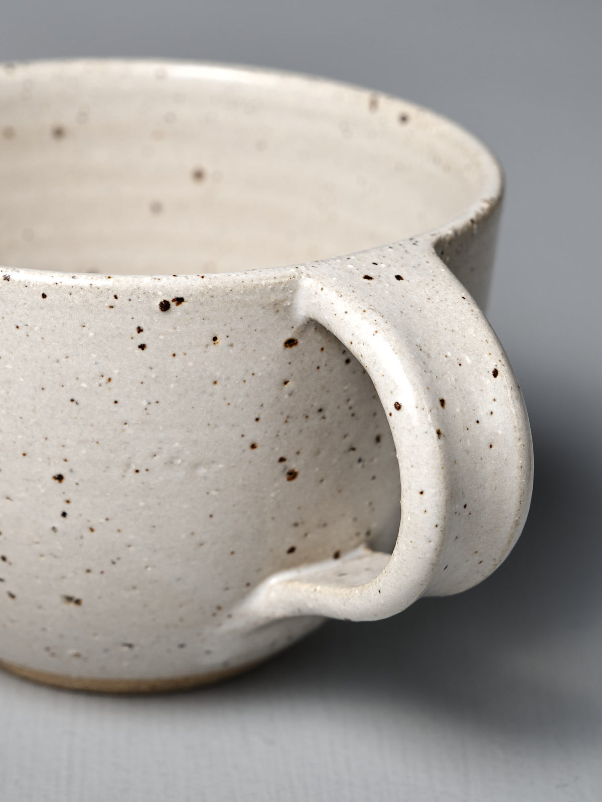 Close-up of the Cup – Speckled White, a handmade piece with satin glaze, curved handle, and speckled white finish, displayed on a plain gray surface.