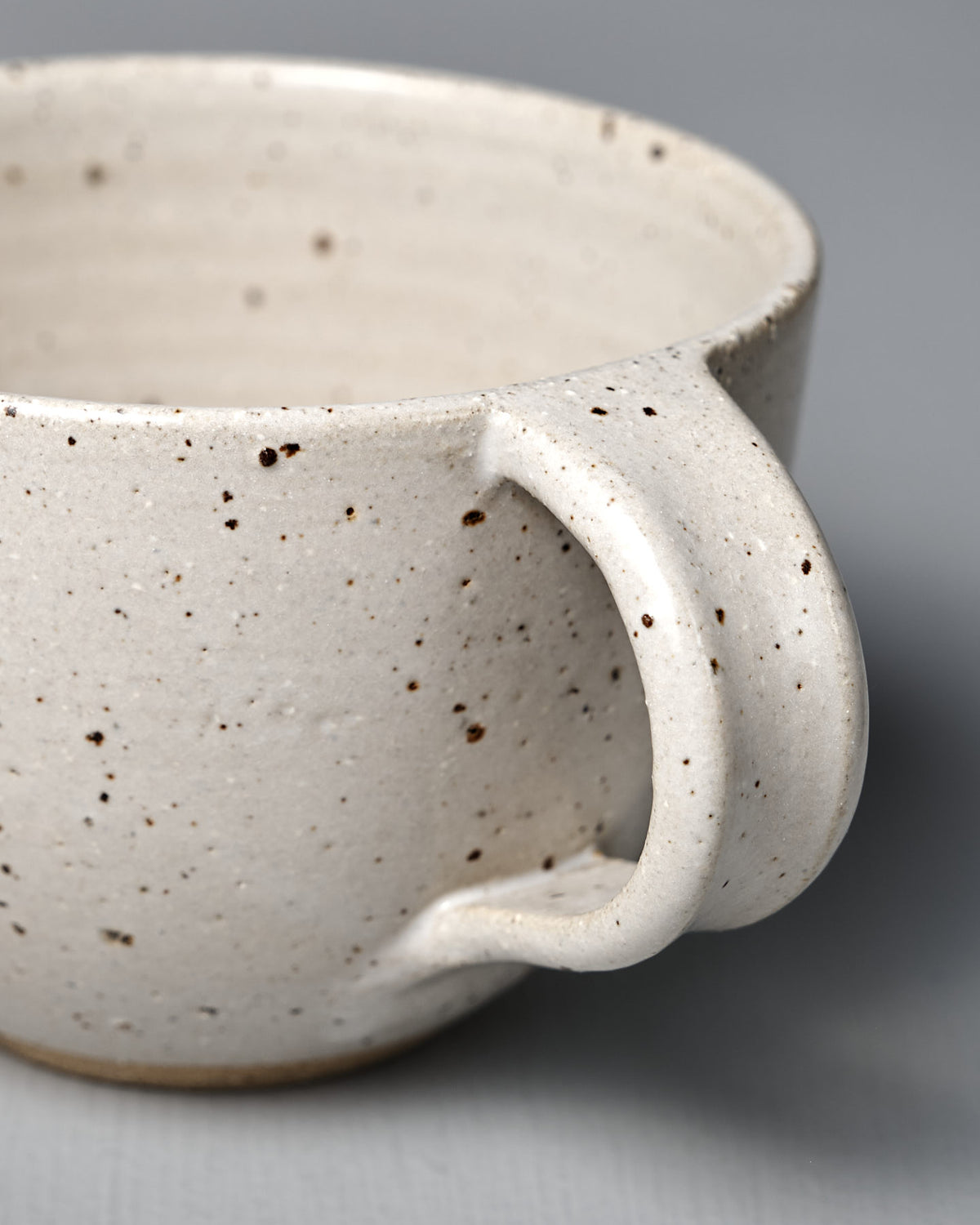 Close-up of the Cup – Speckled White, a handmade piece with satin glaze, curved handle, and speckled white finish, displayed on a plain gray surface.