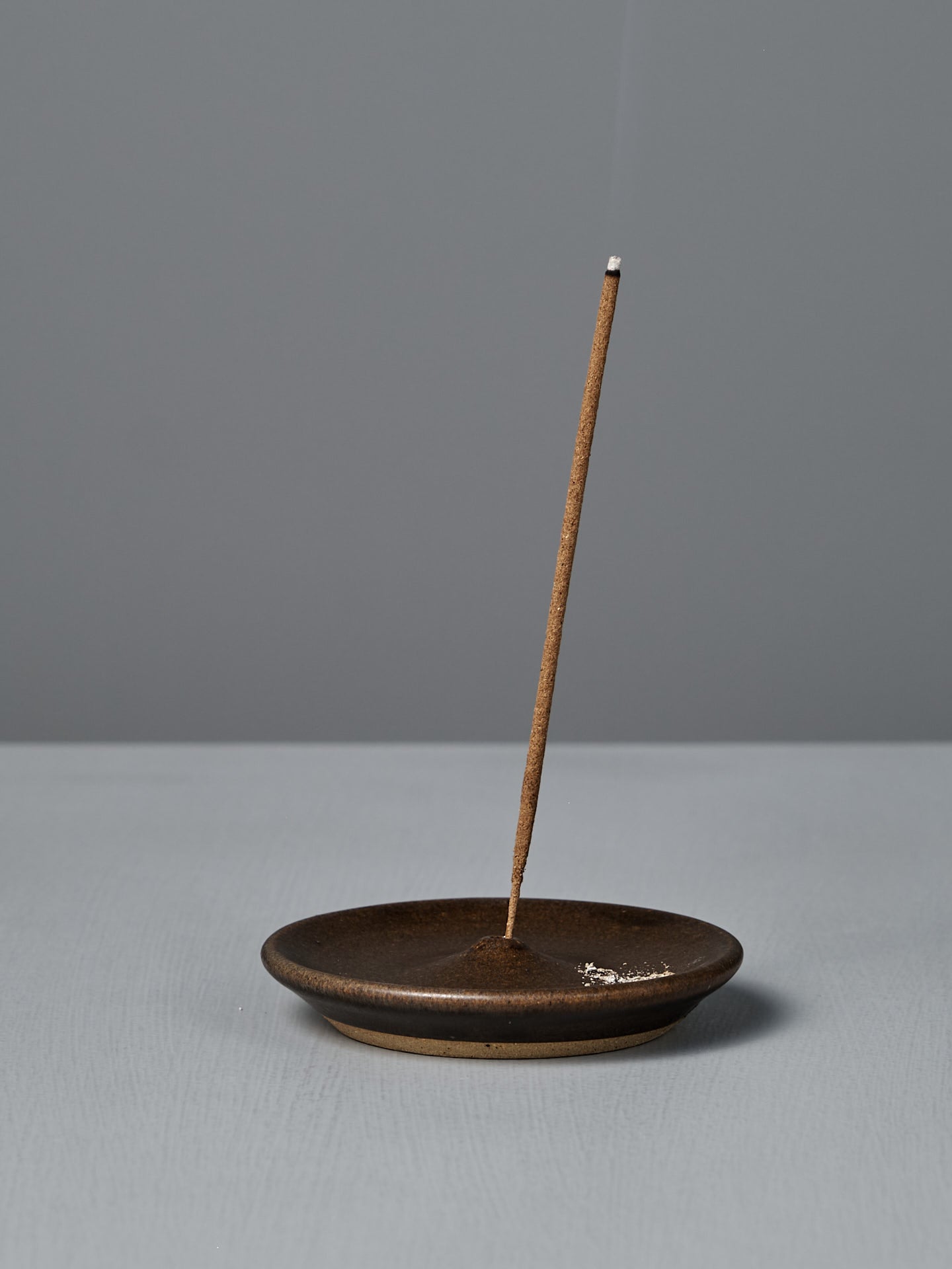 A lit incense stick stands upright in the Incense Holder – Brown on a gray surface, releasing a thin trail of smoke against a plain gray background. Carefully handcrafted in Wellington.