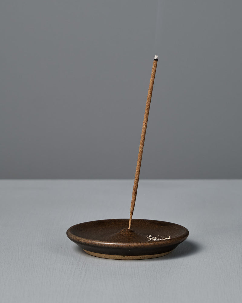 A lit incense stick stands upright in the Incense Holder – Brown on a gray surface, releasing a thin trail of smoke against a plain gray background. Carefully handcrafted in Wellington.