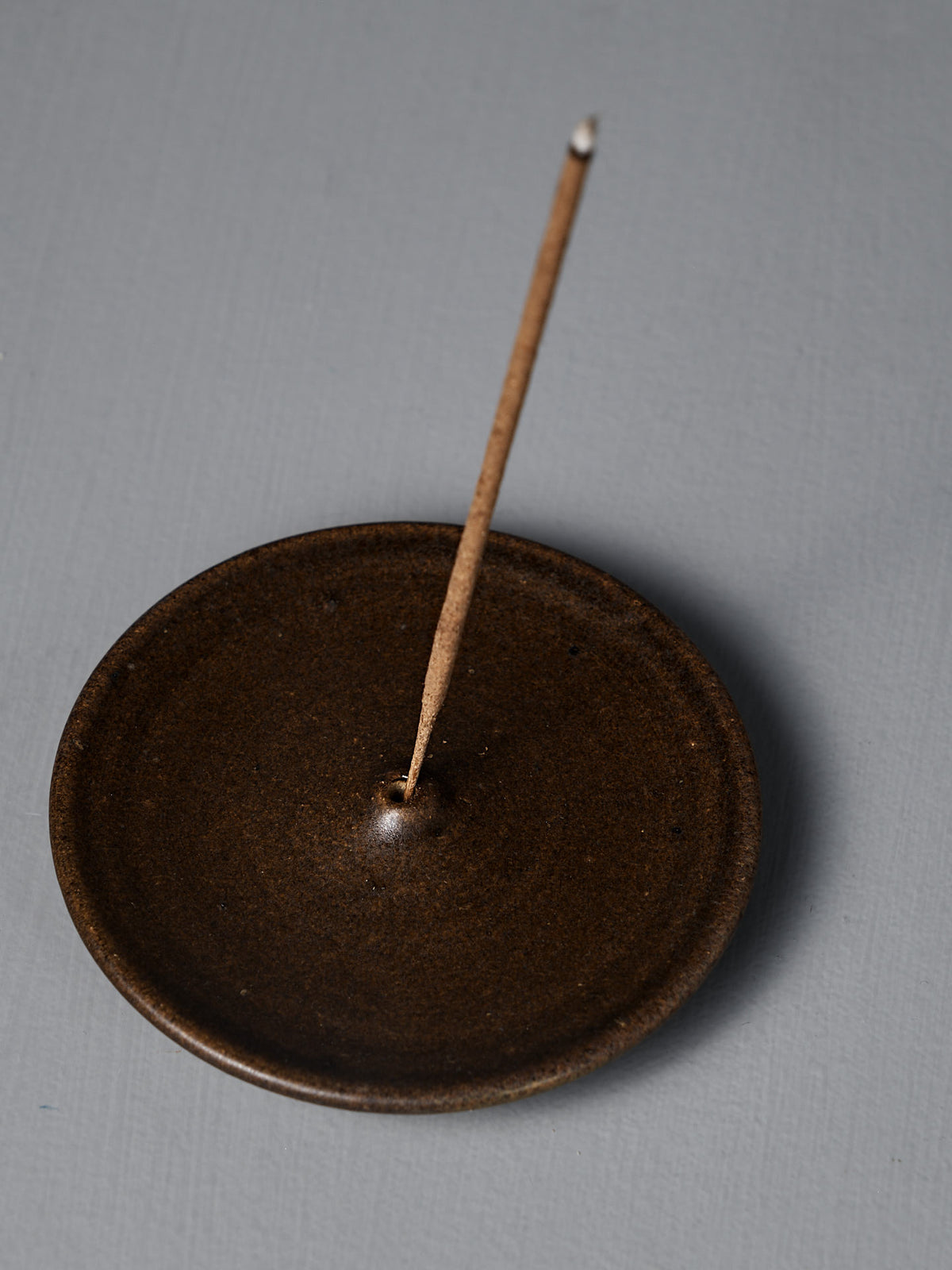 The Incense Holder – Brown is a handmade stoneware piece from Wellington, with a brown ceramic finish, shown holding a single lit incense stick upright on a gray surface.