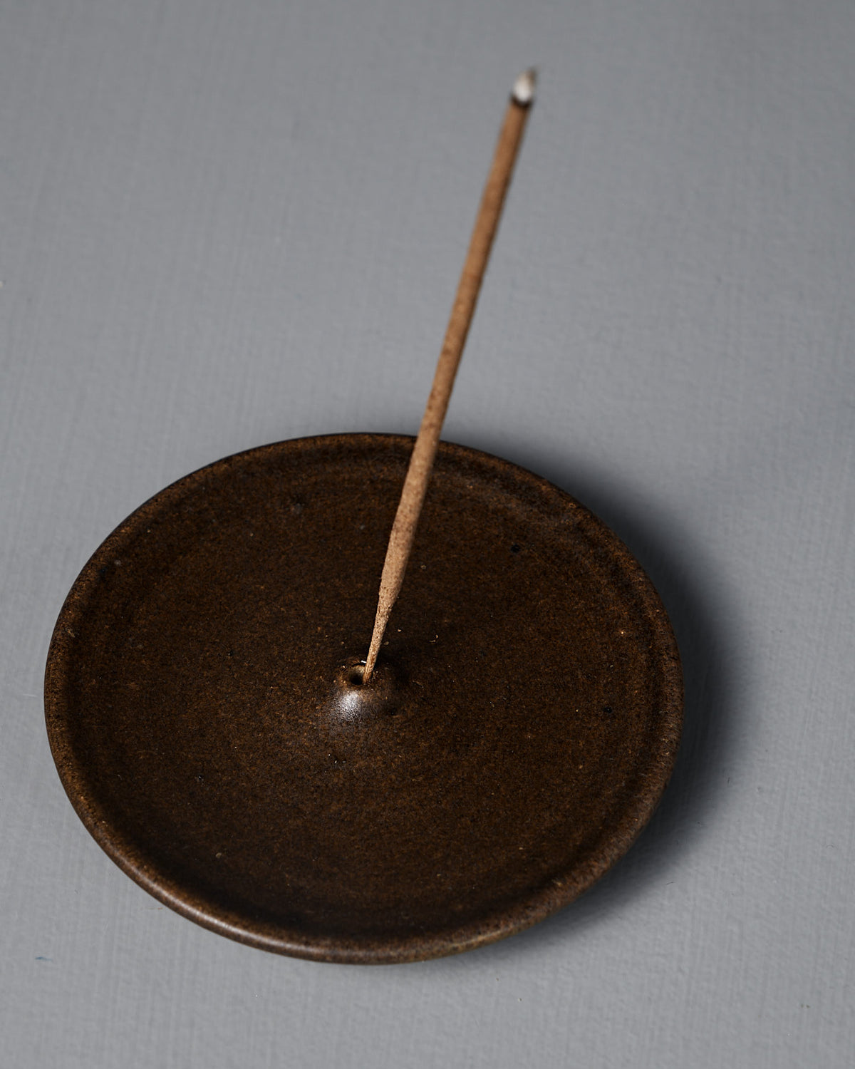 The Incense Holder – Brown is a handmade stoneware piece from Wellington, with a brown ceramic finish, shown holding a single lit incense stick upright on a gray surface.