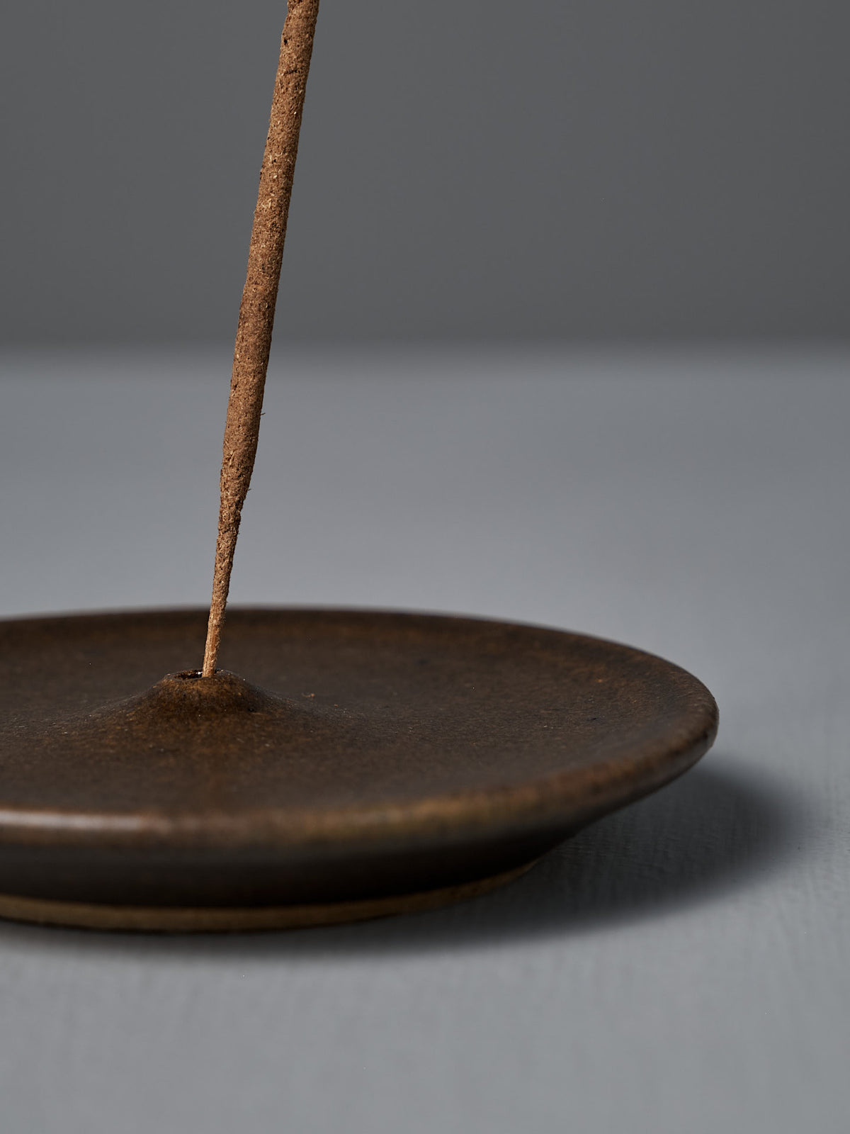 A close-up of the Incense Holder – Brown, a handmade stoneware stick incense holder crafted in Wellington, shown with a brown incense stick on a gray surface and background.