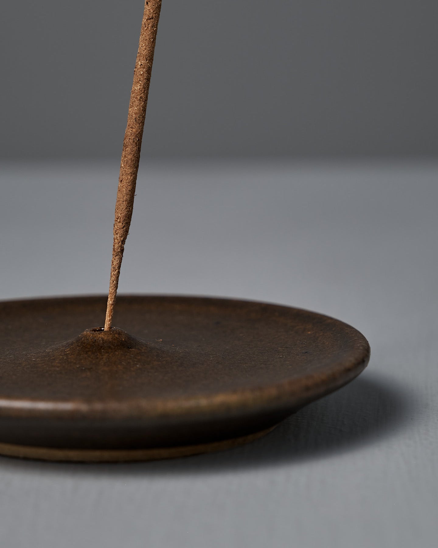 A lit incense stick stands upright in the Incense Holder – Brown on a gray surface, releasing a thin trail of smoke against a plain gray background. Carefully handcrafted in Wellington.
