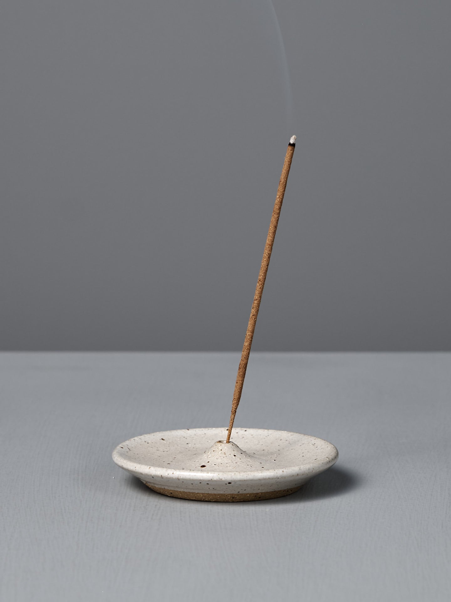 A lit incense stick stands upright in the Incense Holder – Speckled White, with a thin trail of smoke rising against a plain gray background.