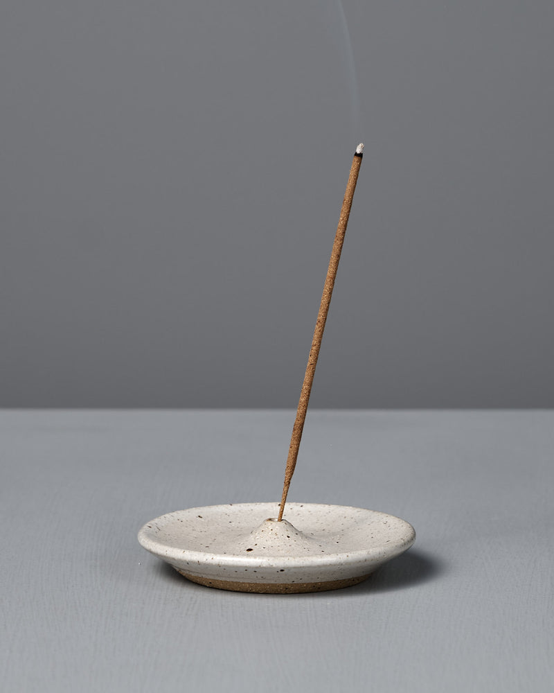 A lit incense stick stands upright in the Incense Holder – Speckled White, with a thin trail of smoke rising against a plain gray background.