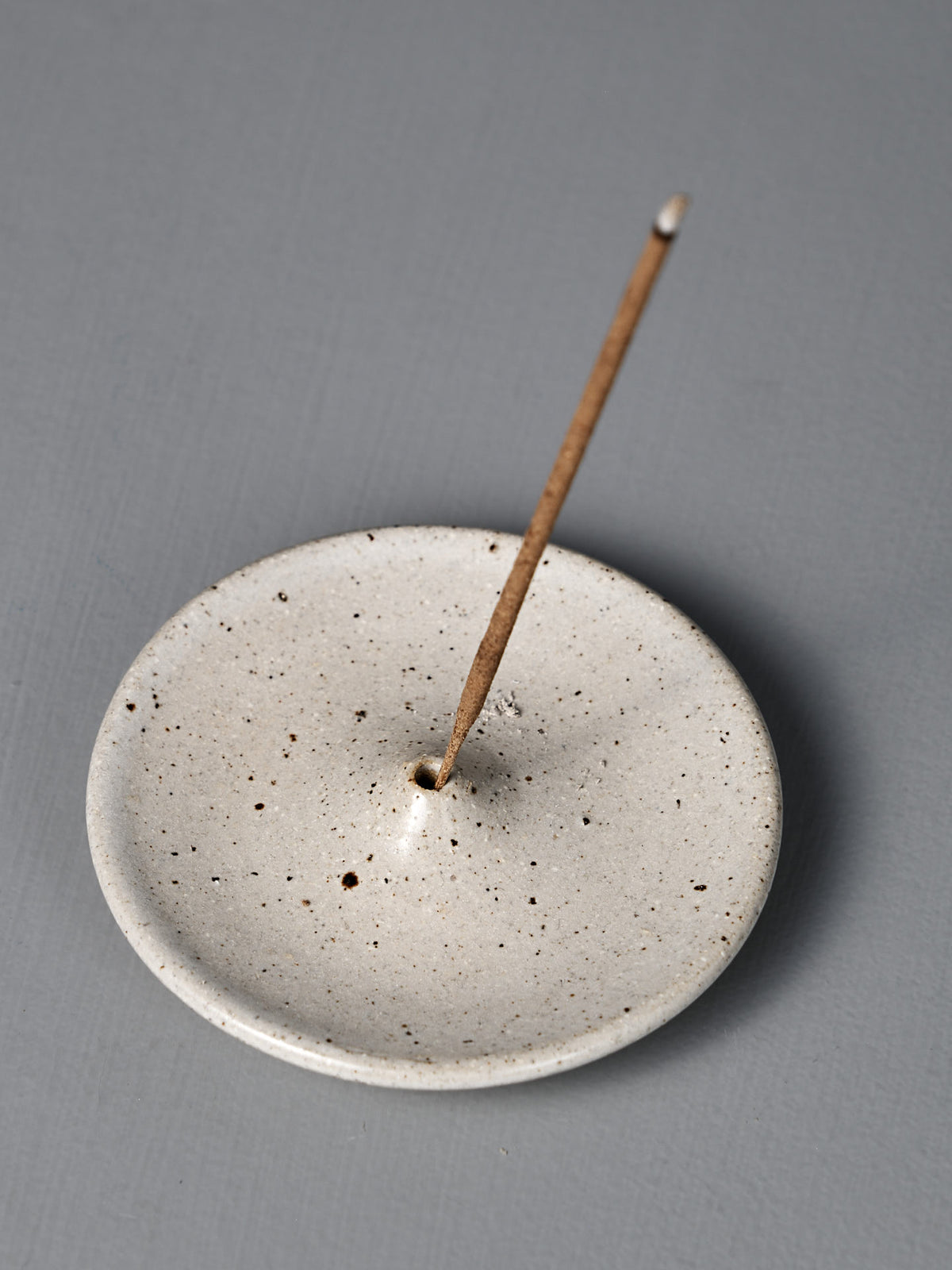 The Incense Holder – Speckled White, a handmade stoneware piece from Pōneke Wellington, displays its unique speckled finish while holding a single lit incense stick on a gray surface.